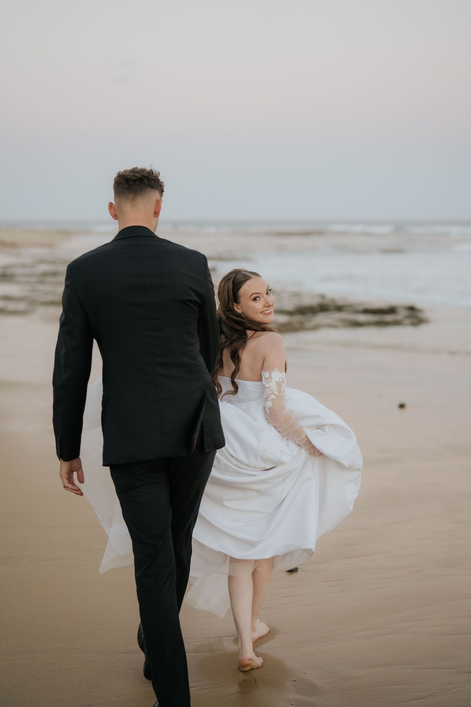 Bride in white off-shoulder gown walks barefoot on beach with groom in black suit, she glances back smiling.