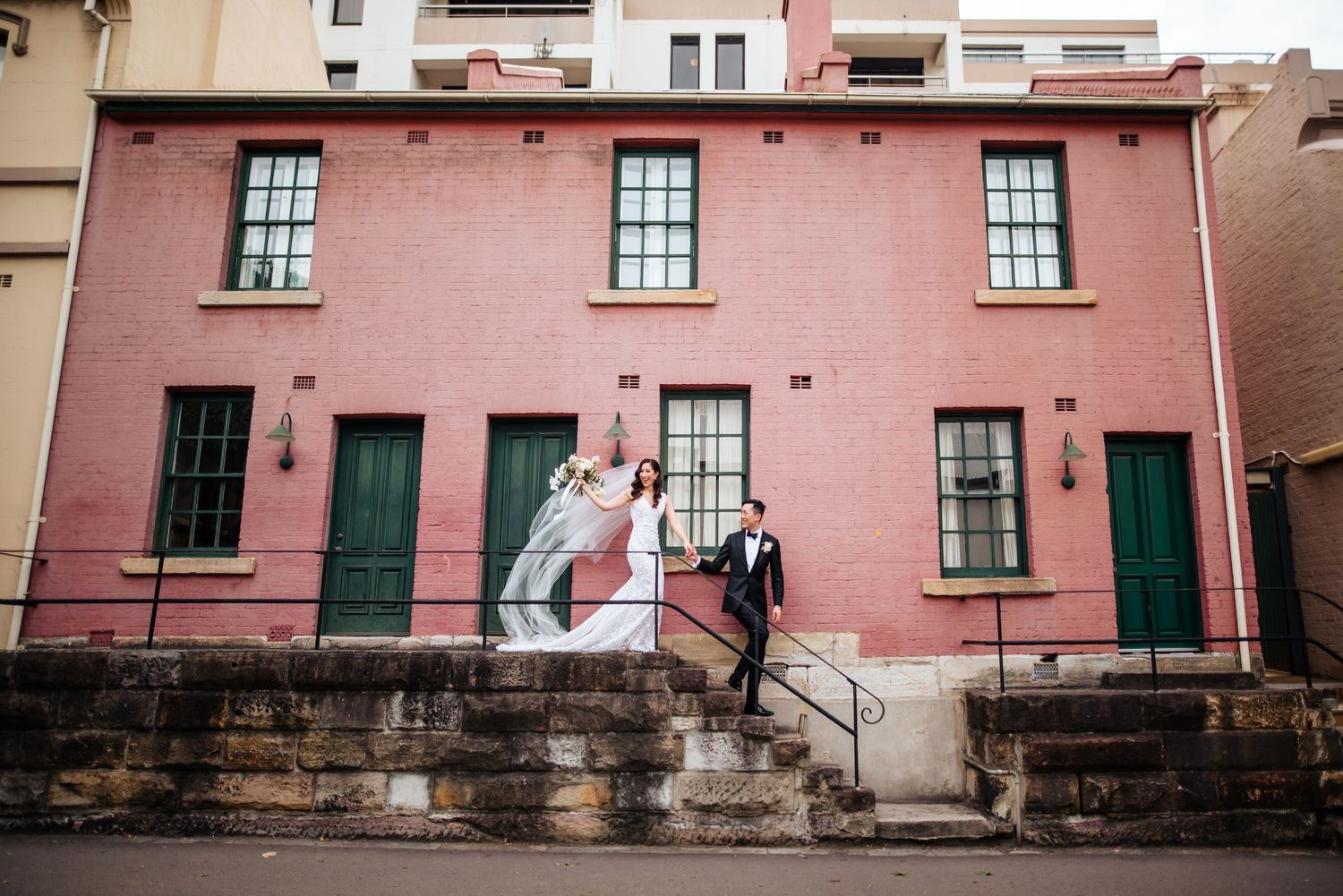 Bride and groom posing on stone steps outside a charming pink building with green doors in Sydney.