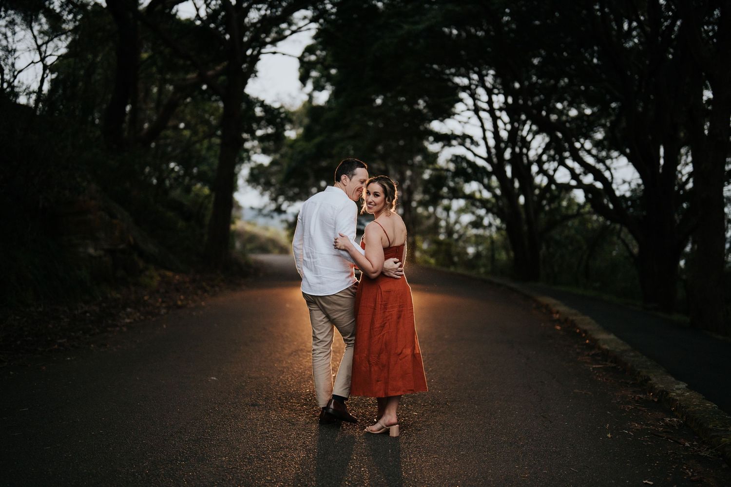 Couple shares a tender embrace on a tree-lined road at dusk during a romantic photo session.