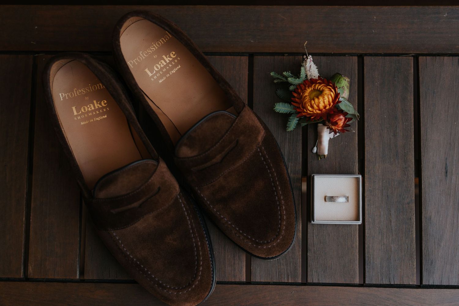 Loake brown suede penny loafers beside a floral boutonniere and wedding ring on dark wood floor.