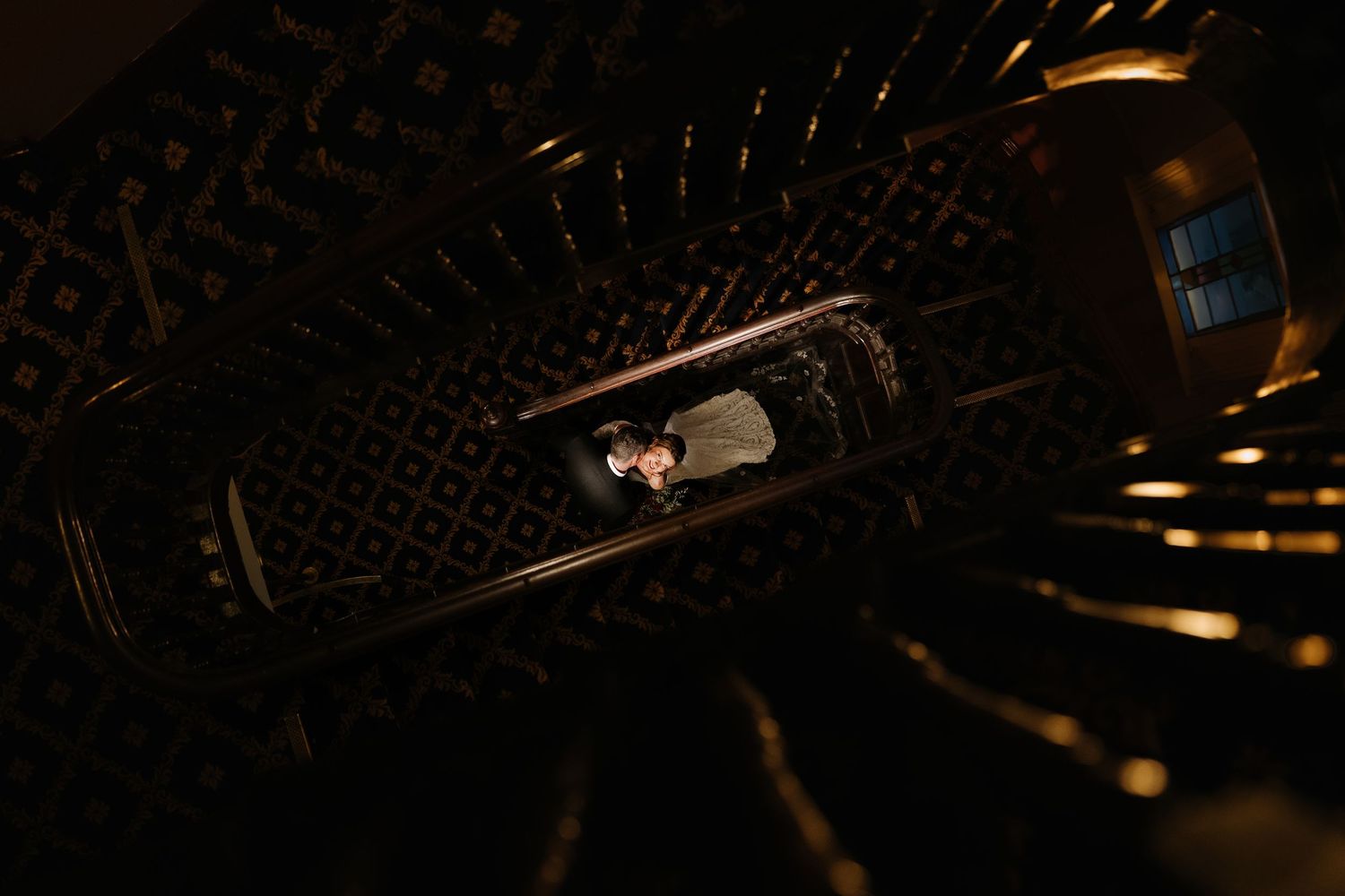 Aerial view of a couple standing in a dark spiral staircase, illuminated by warm golden light.