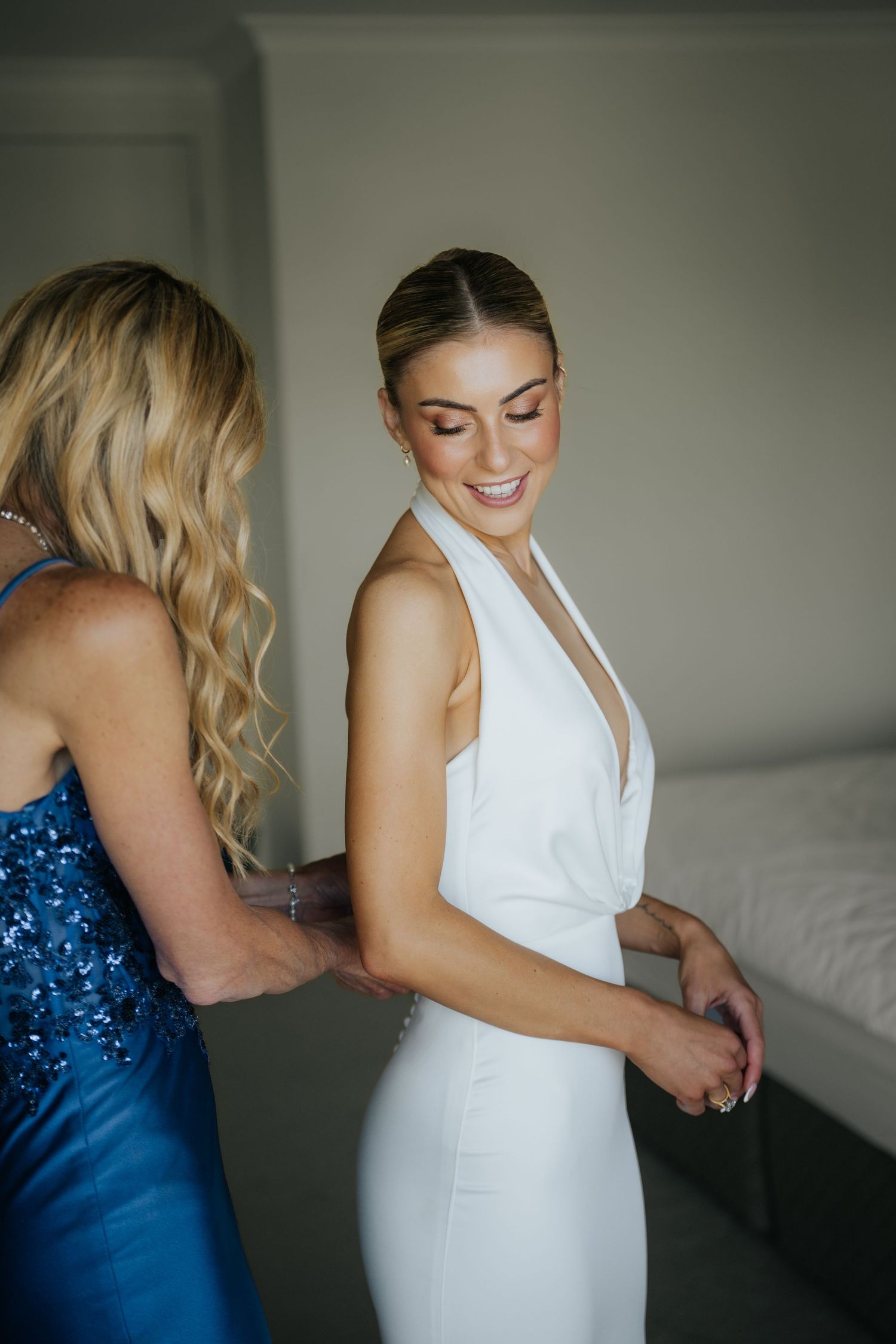 Smiling bride in white halter wedding dress being helped by woman in blue sequin gown.