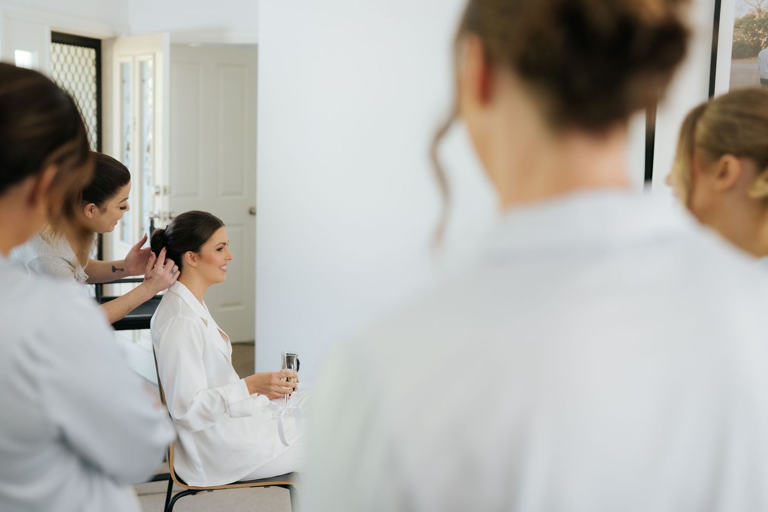 Bridesmaids in white robes help a bride get ready for her wedding day in a bright, elegant room.