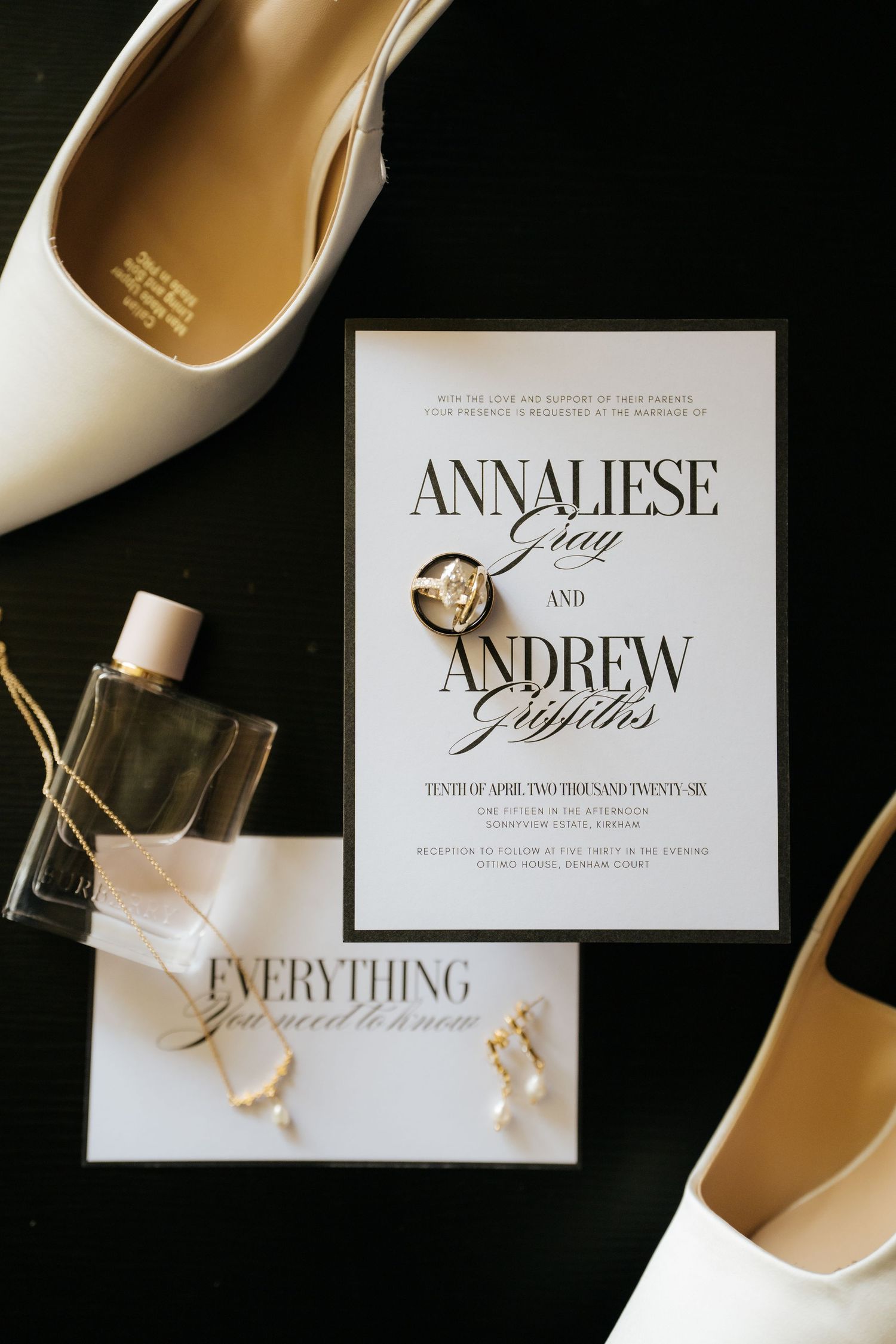 Wedding flat lay featuring Annaliese and Andrew invitation, diamond ring, pearl earrings, white heels, and perfume on dark surface.