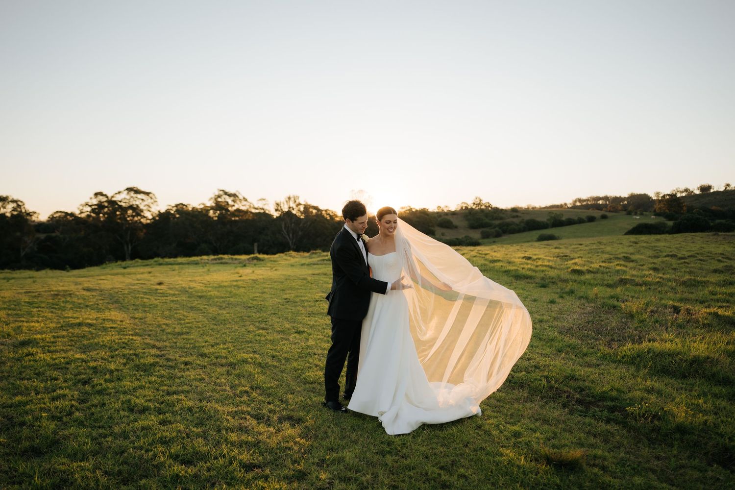 Bride and groom embrace at golden sunset in green field, her long veil flowing in the breeze.