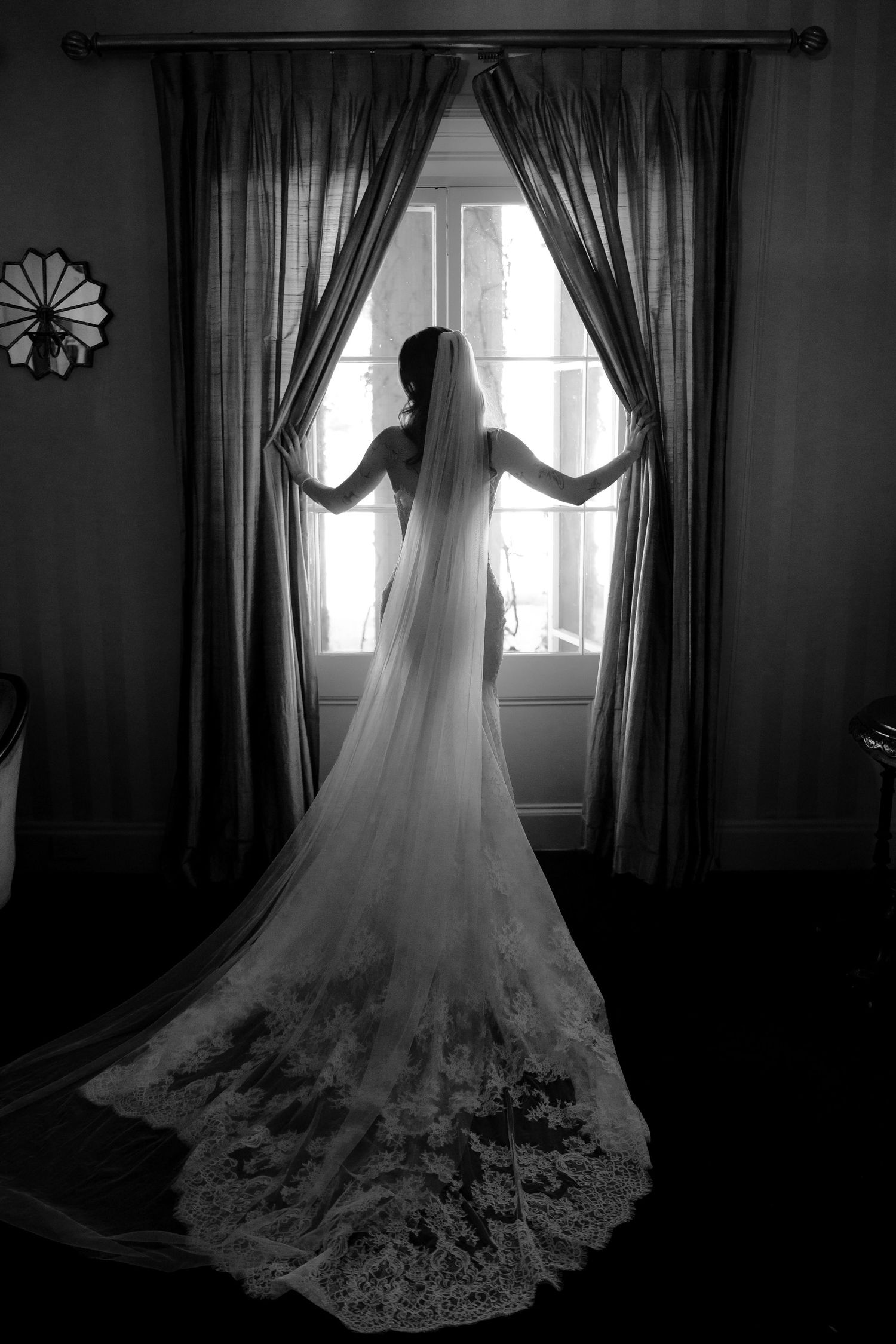 Bride in a lace-trimmed cathedral veil stands silhouetted before a draped window in black and white photo.