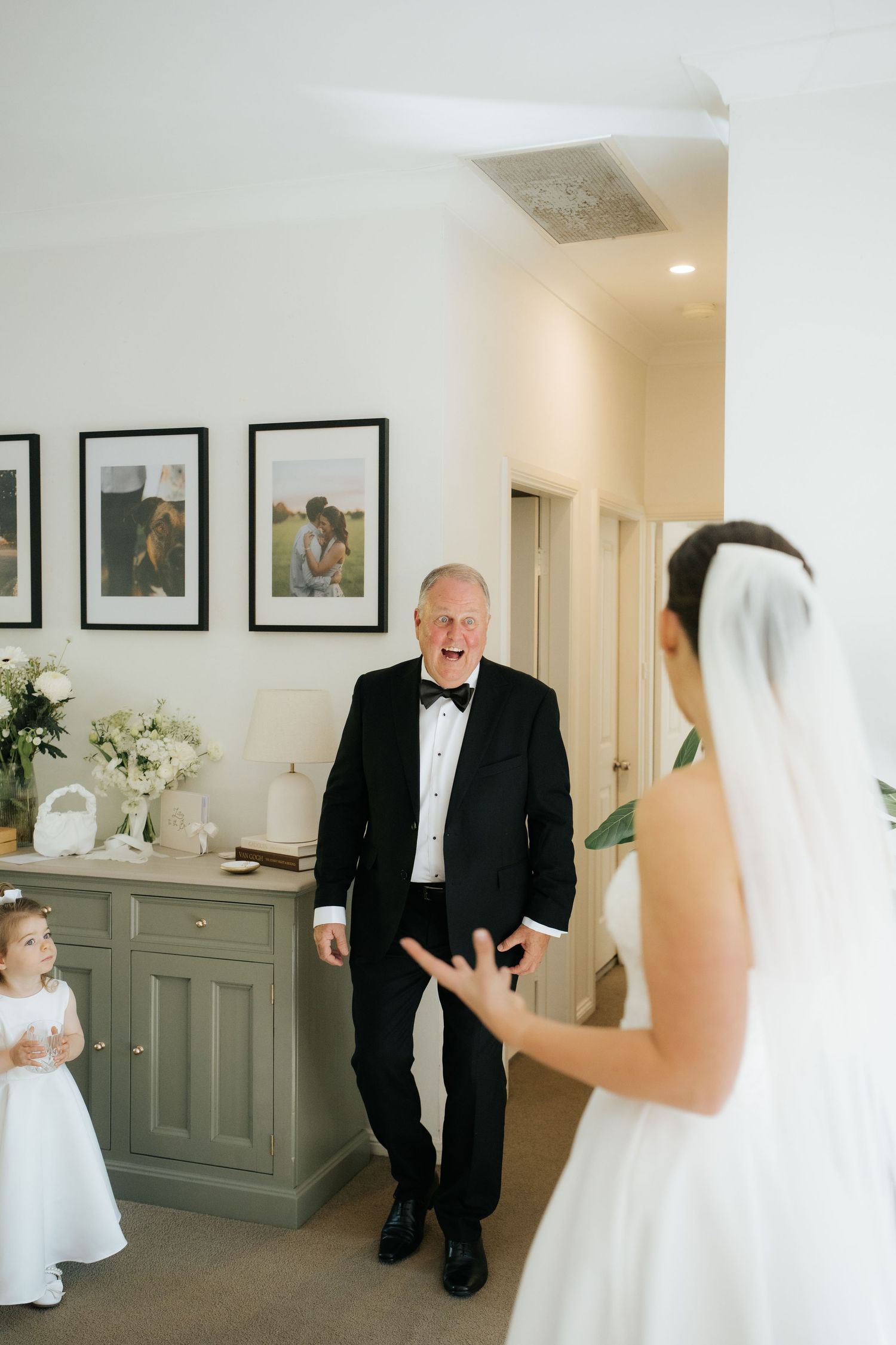 Emotional father reacting with joy seeing bride in wedding dress during first look moment indoors.