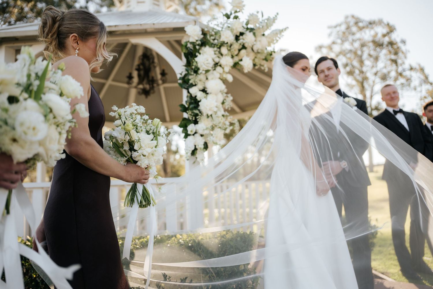 Bride in white gown with flowing veil at outdoor wedding ceremony with floral arch.