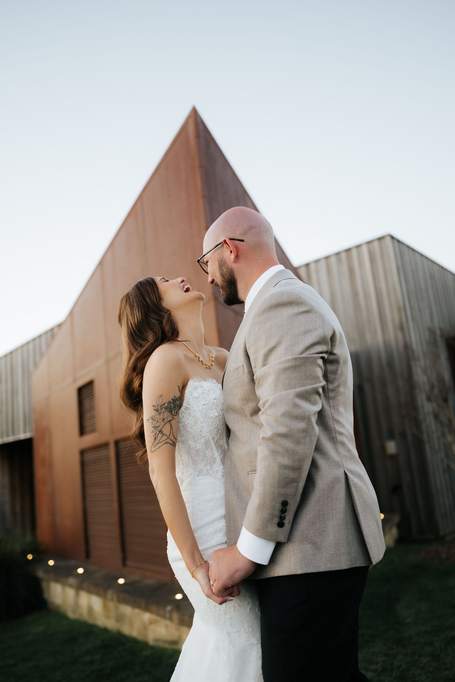Bride and groom holding hands and gazing at each other in front of a modern copper building at dusk.