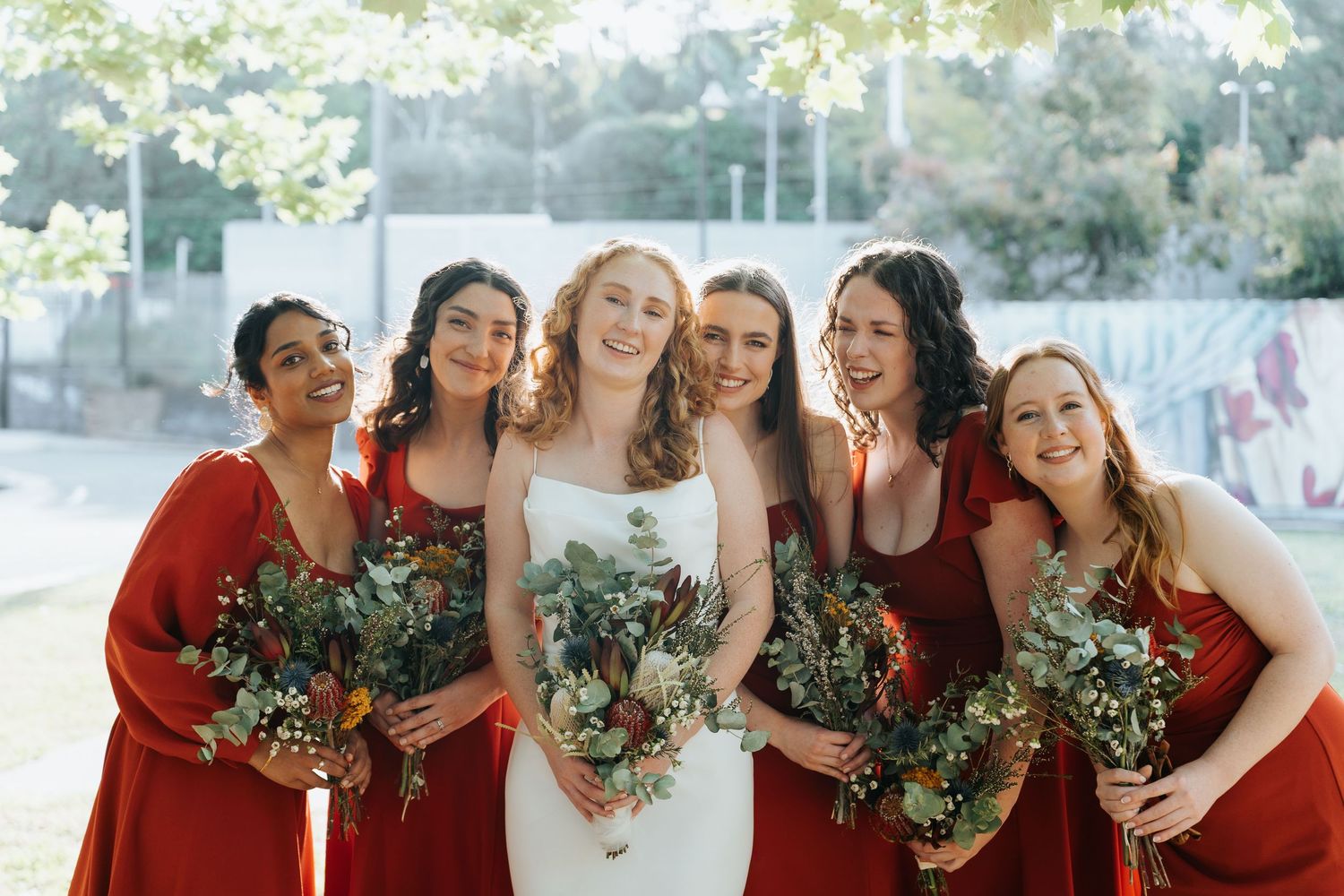 Bride in white gown with five bridesmaids in red dresses holding green bouquets, smiling outdoors.