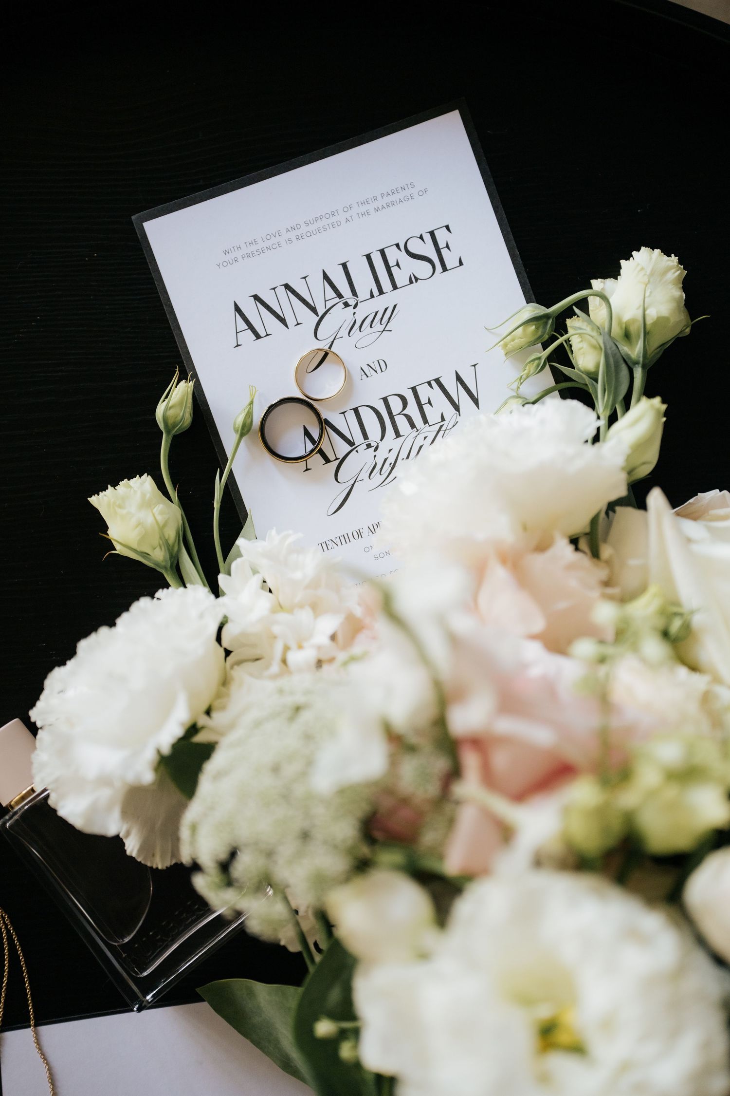 Wedding invitation for Annaliese Gray and Andrew displayed with gold rings on a white floral bouquet of roses and lisianthus.