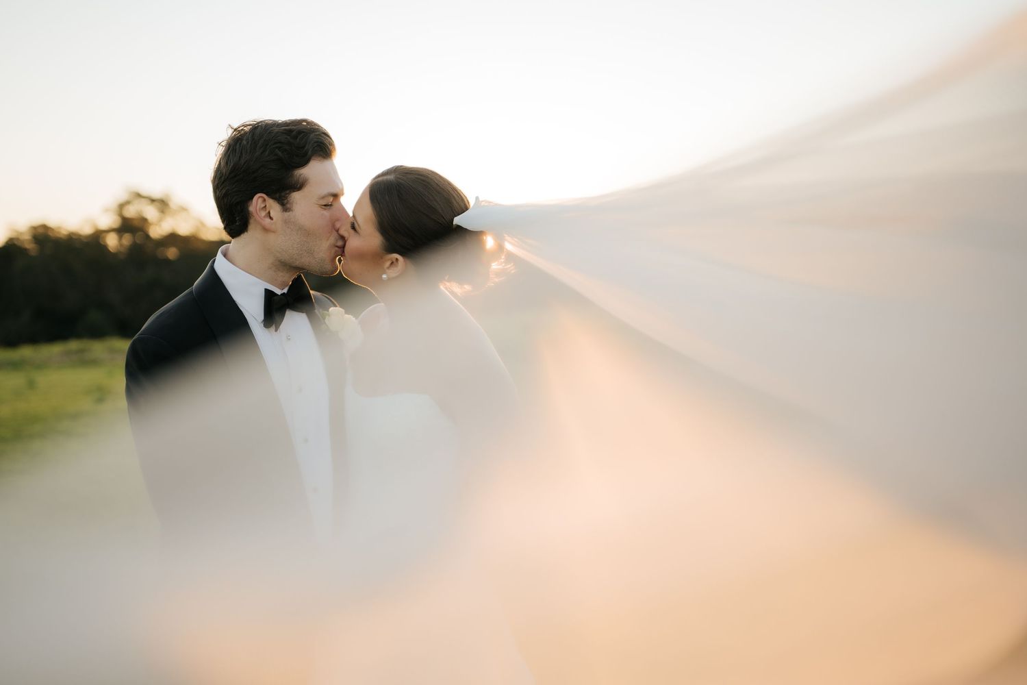 Bride and groom sharing a kiss at sunset with veil flowing in the wind.