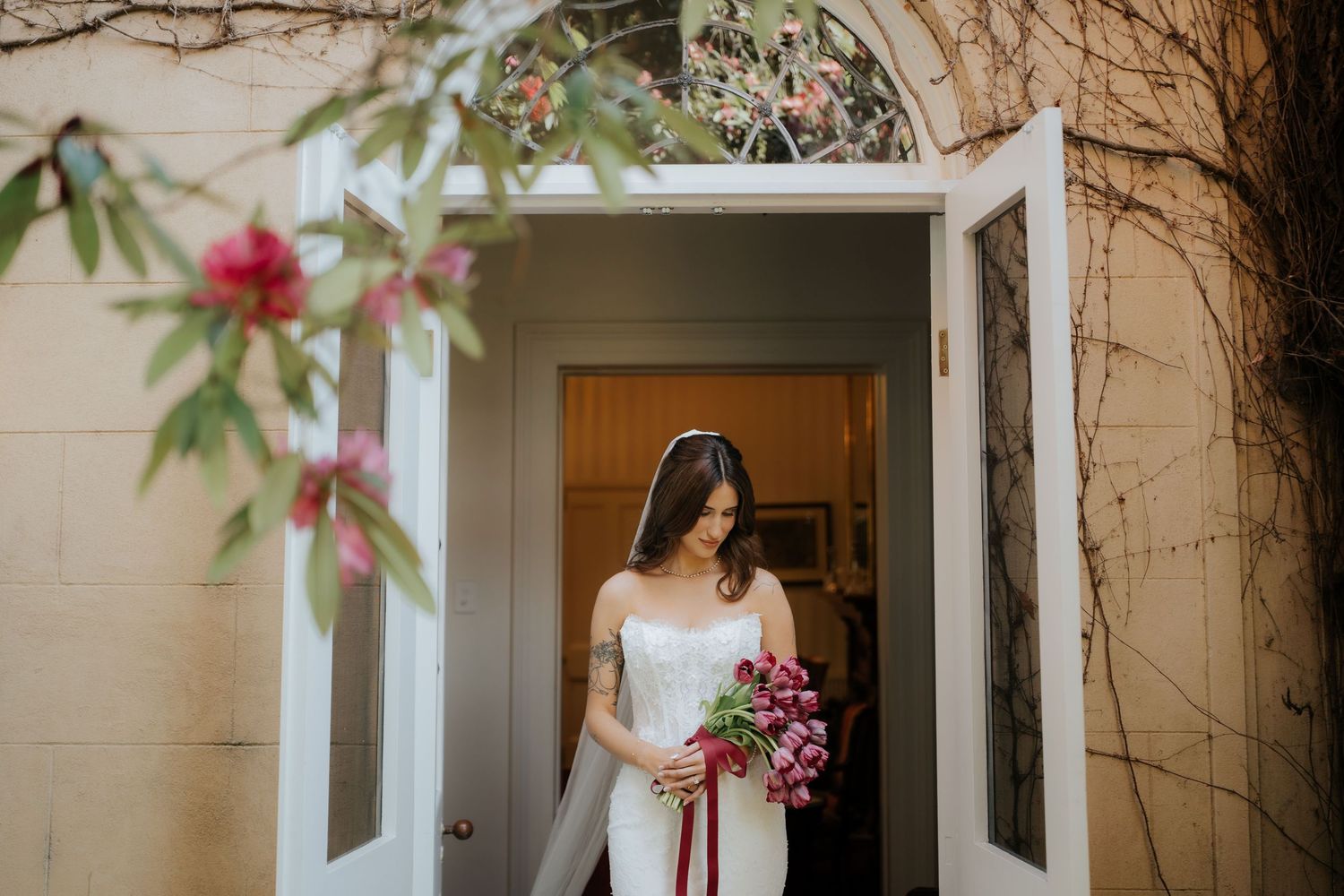 Bride in white gown holding red bouquet stands in doorway surrounded by pink blooms.