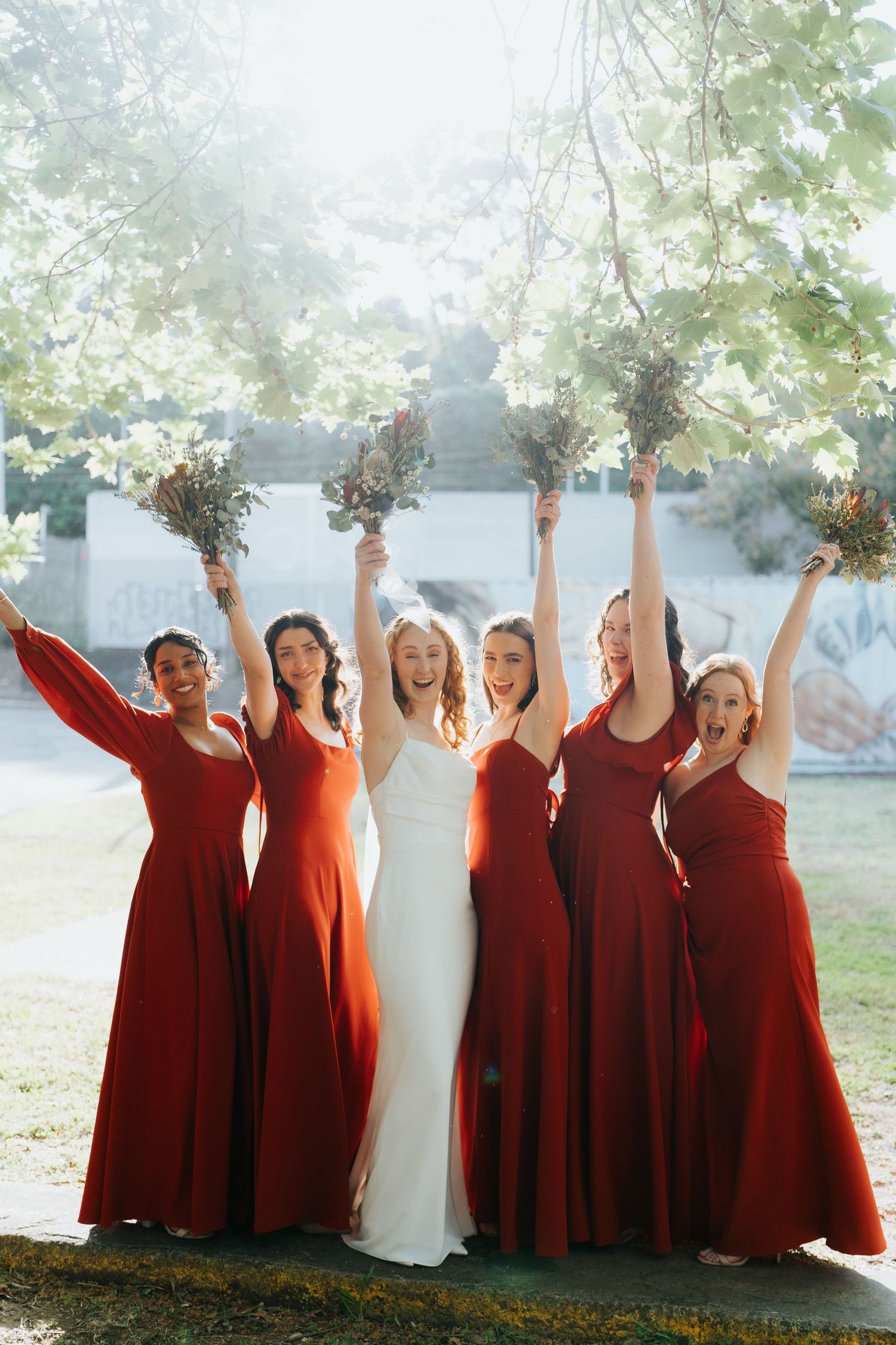 Bride in white gown with five bridesmaids in red dresses raising bouquets outdoors under blooming trees.