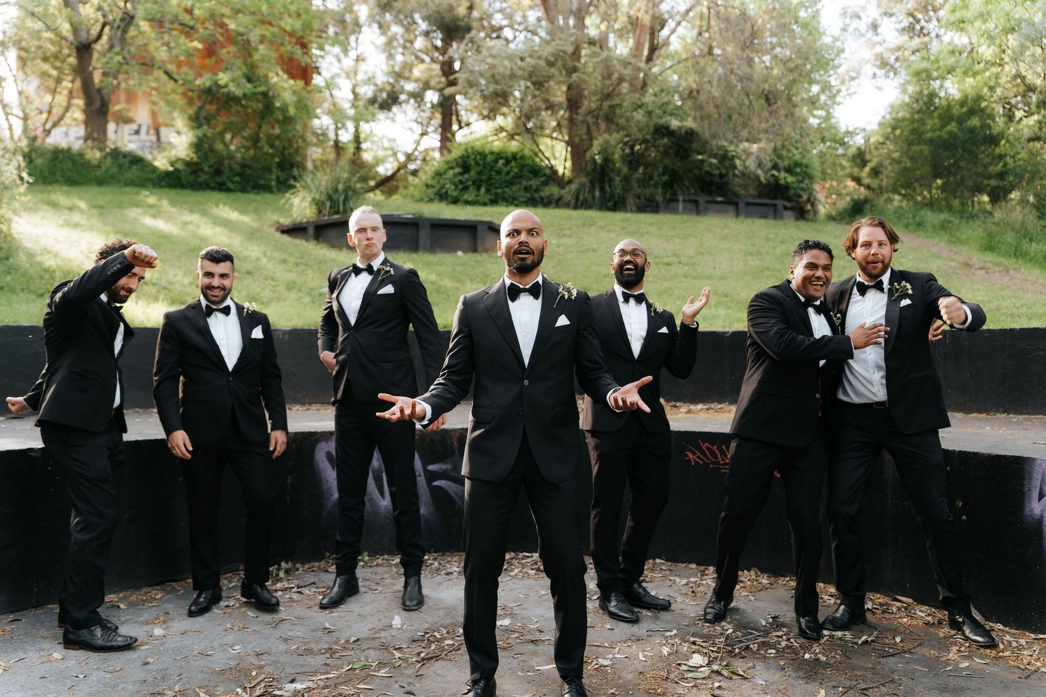 Groom and groomsmen in black tuxedos posing outdoors near a stone wall with trees in background.