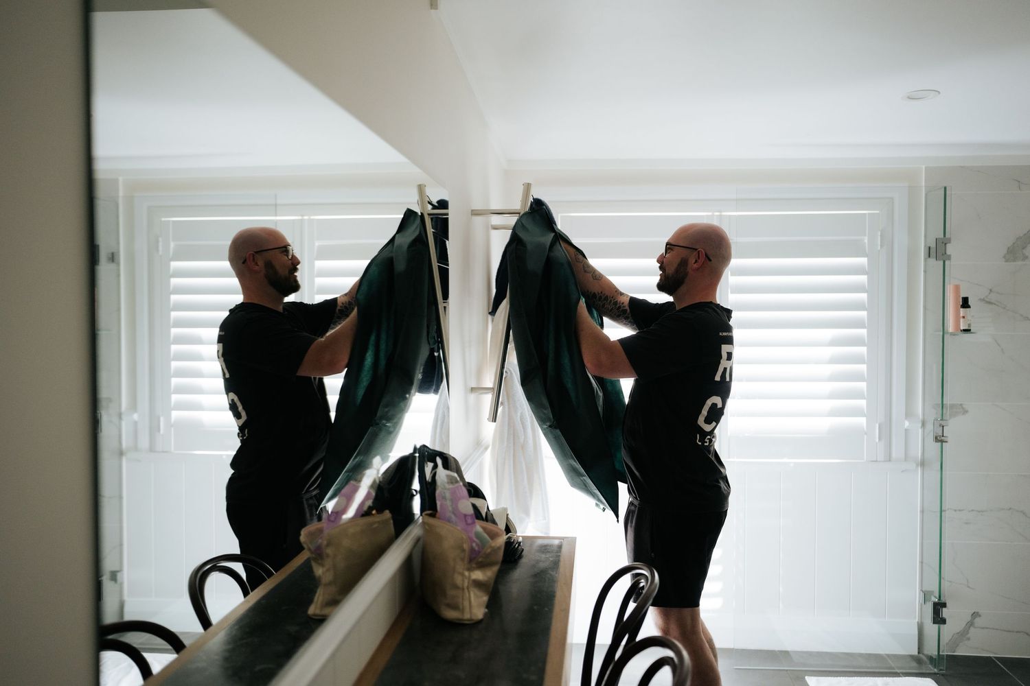 Bald man holding dark wetsuit reflected in bathroom mirror with white shuttered windows in background.