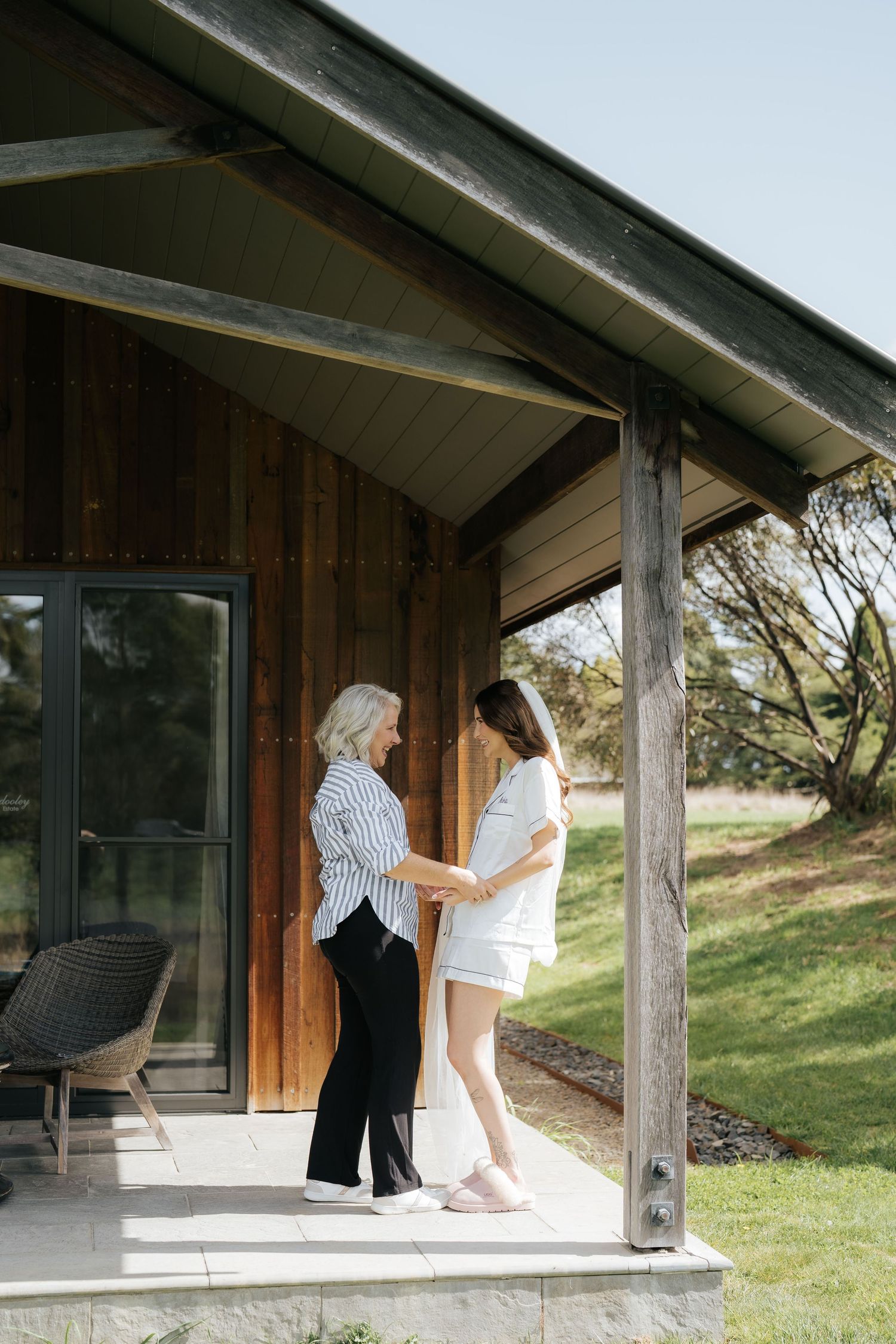 Bride in white dress sharing tender moment with older woman on rustic wooden cabin porch outdoors.