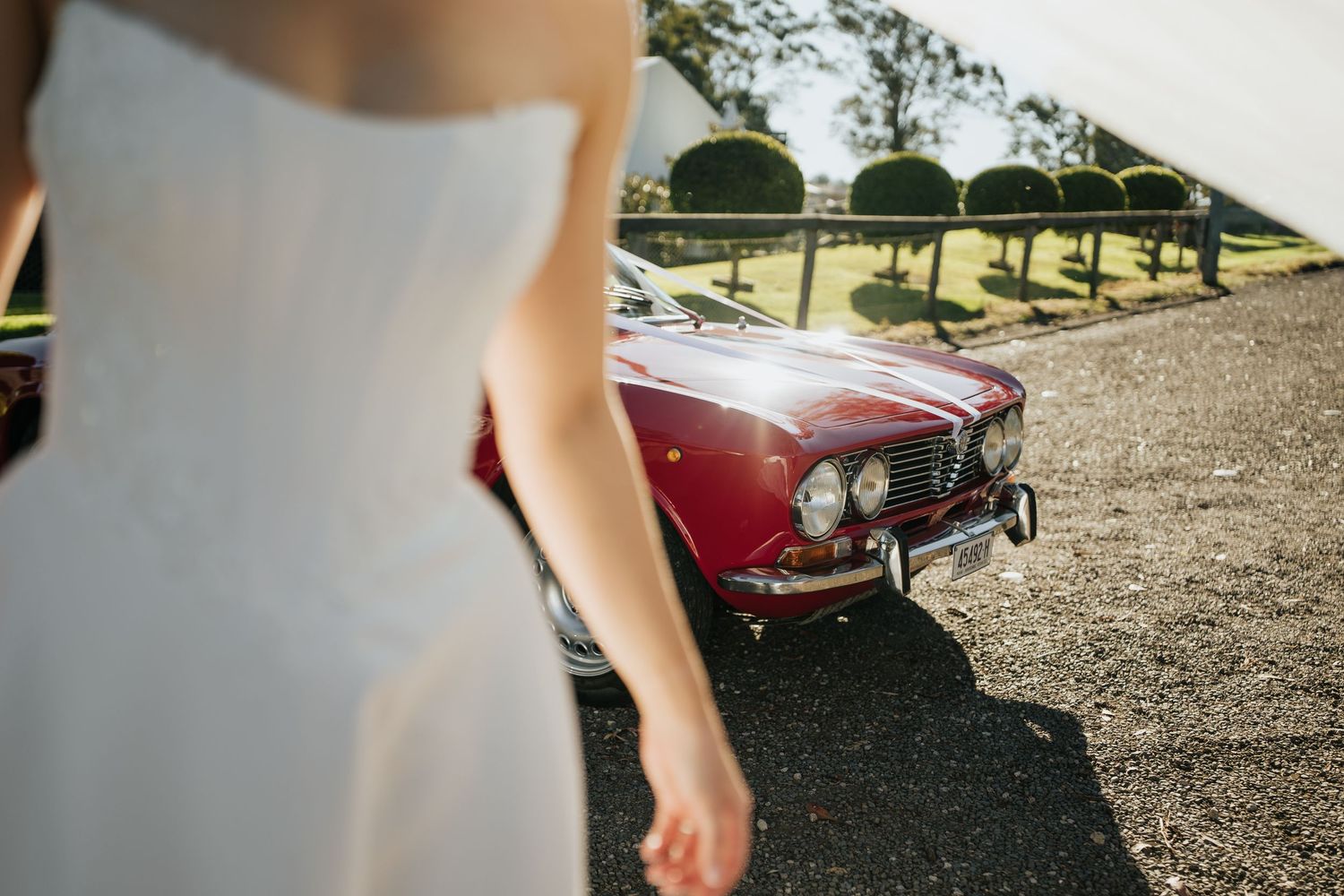 Bride in white wedding dress walking beside a classic red vintage car on a sunny day.