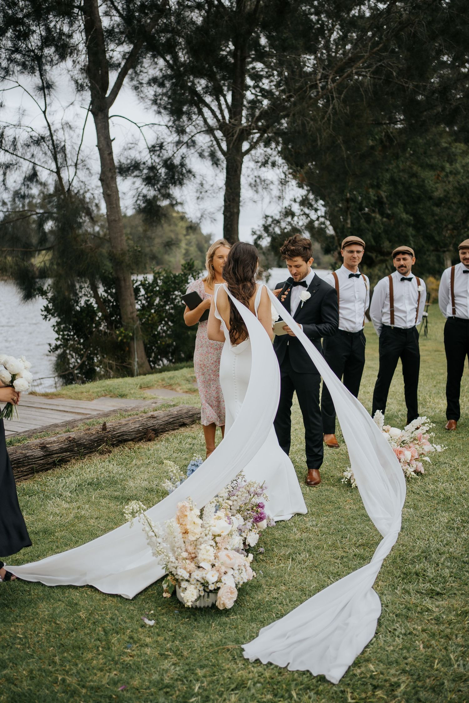 Bride in flowing white gown with long veil stands at outdoor waterside ceremony with wedding party.