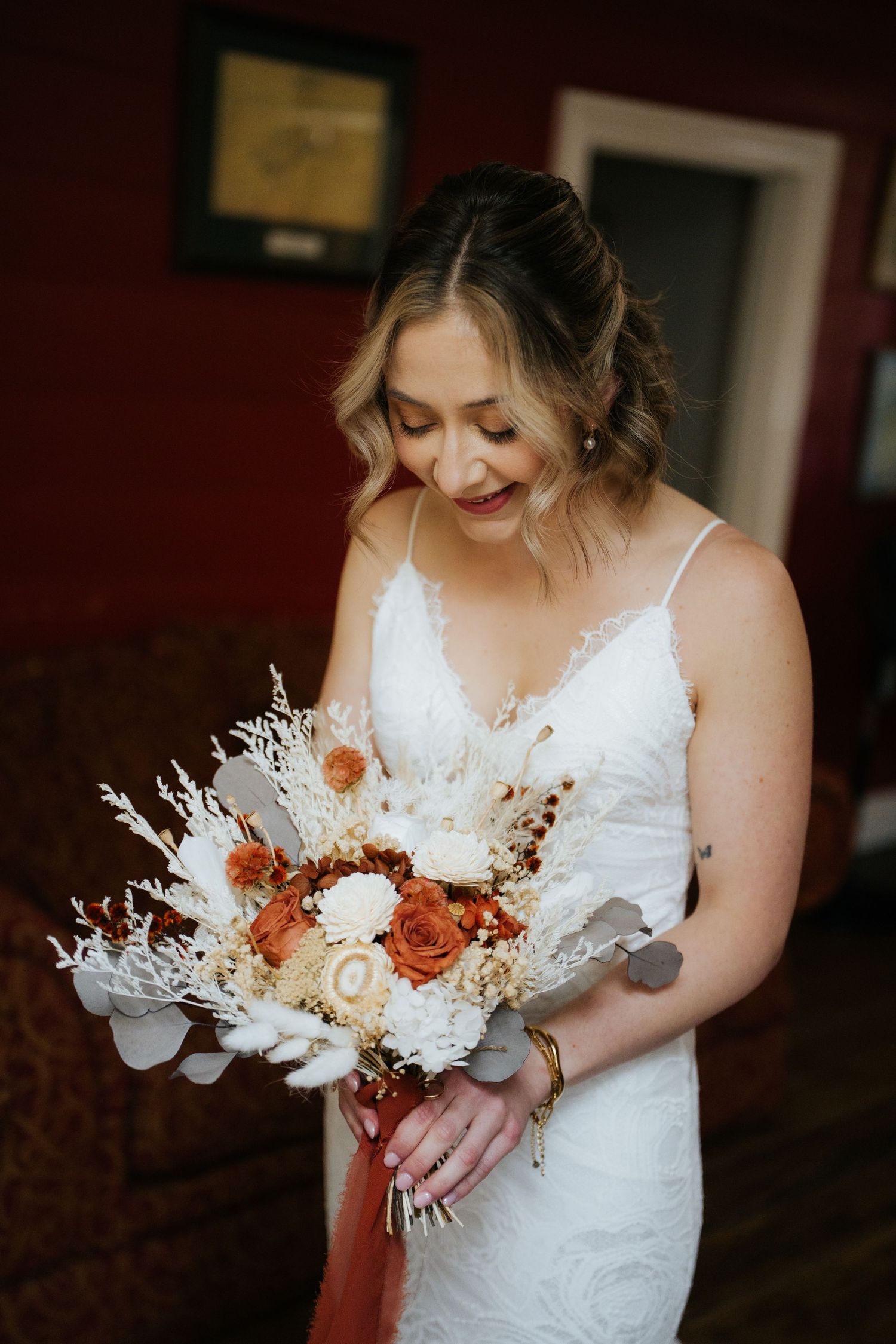 Bride in white lace gown holding a rustic bouquet of orange and ivory dried flowers, smiling softly indoors.