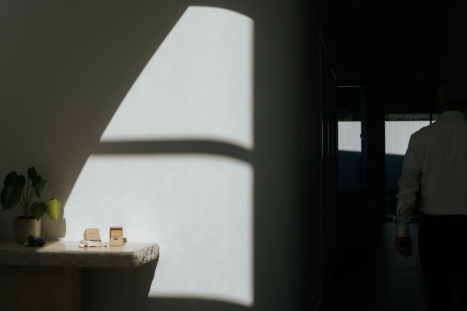 Dramatic sunlight casting geometric shadows on a minimalist white room with a small table, plant, and person standing in a doorway.