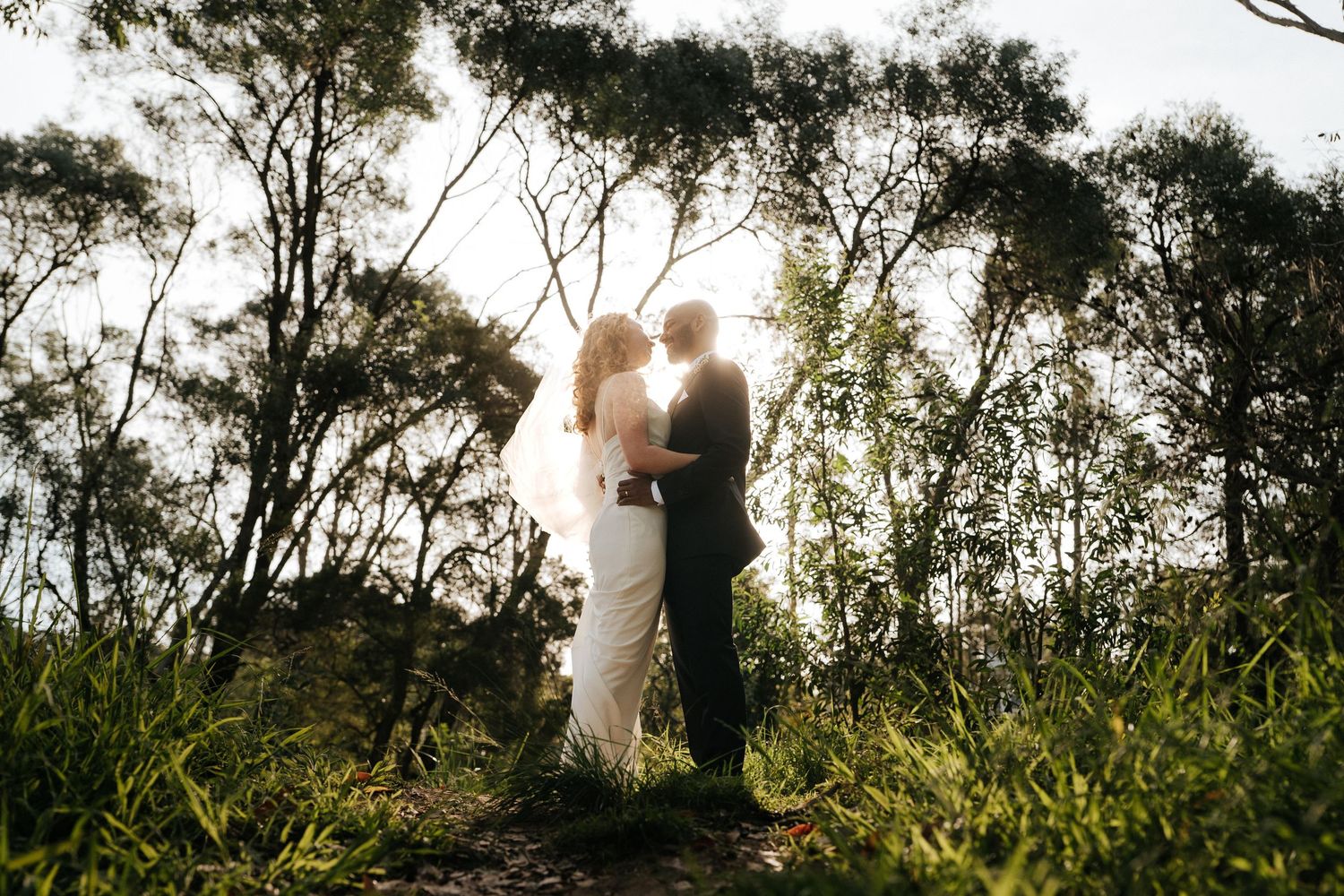 Bride and groom silhouetted against golden sunlight in a lush forest setting during their outdoor wedding ceremony.