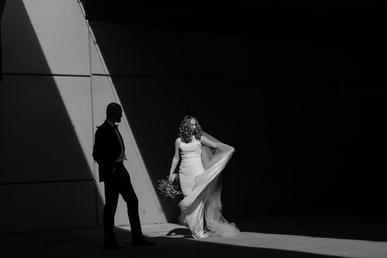 Black and white wedding photo of bride and groom in dramatic light and shadow against a modern wall.