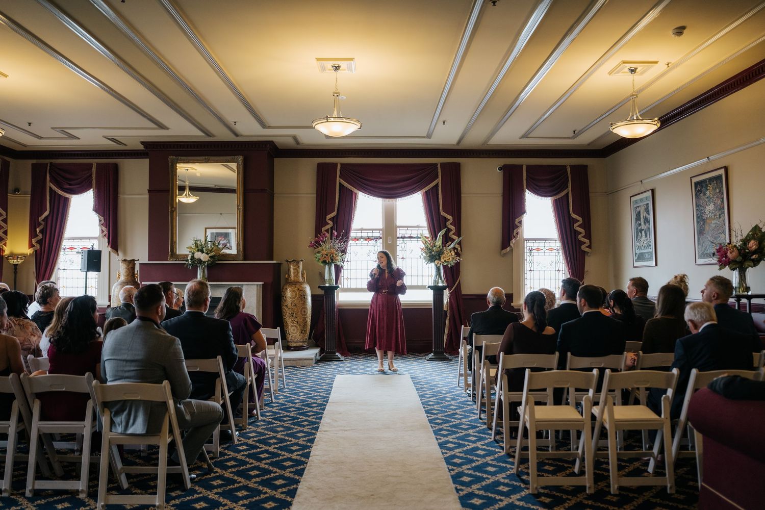 Elegant indoor wedding ceremony with guests seated in rows facing officiant in burgundy dress, white aisle runner, classic decor.