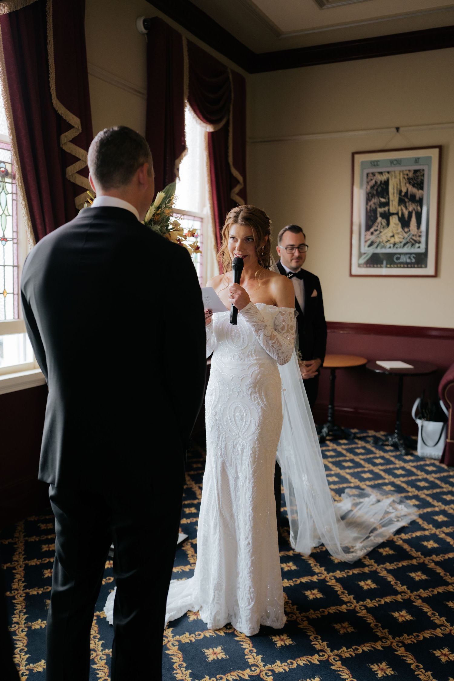 Bride in lace off-shoulder gown recites vows holding microphone, groom in black suit, officiant behind in elegant venue.