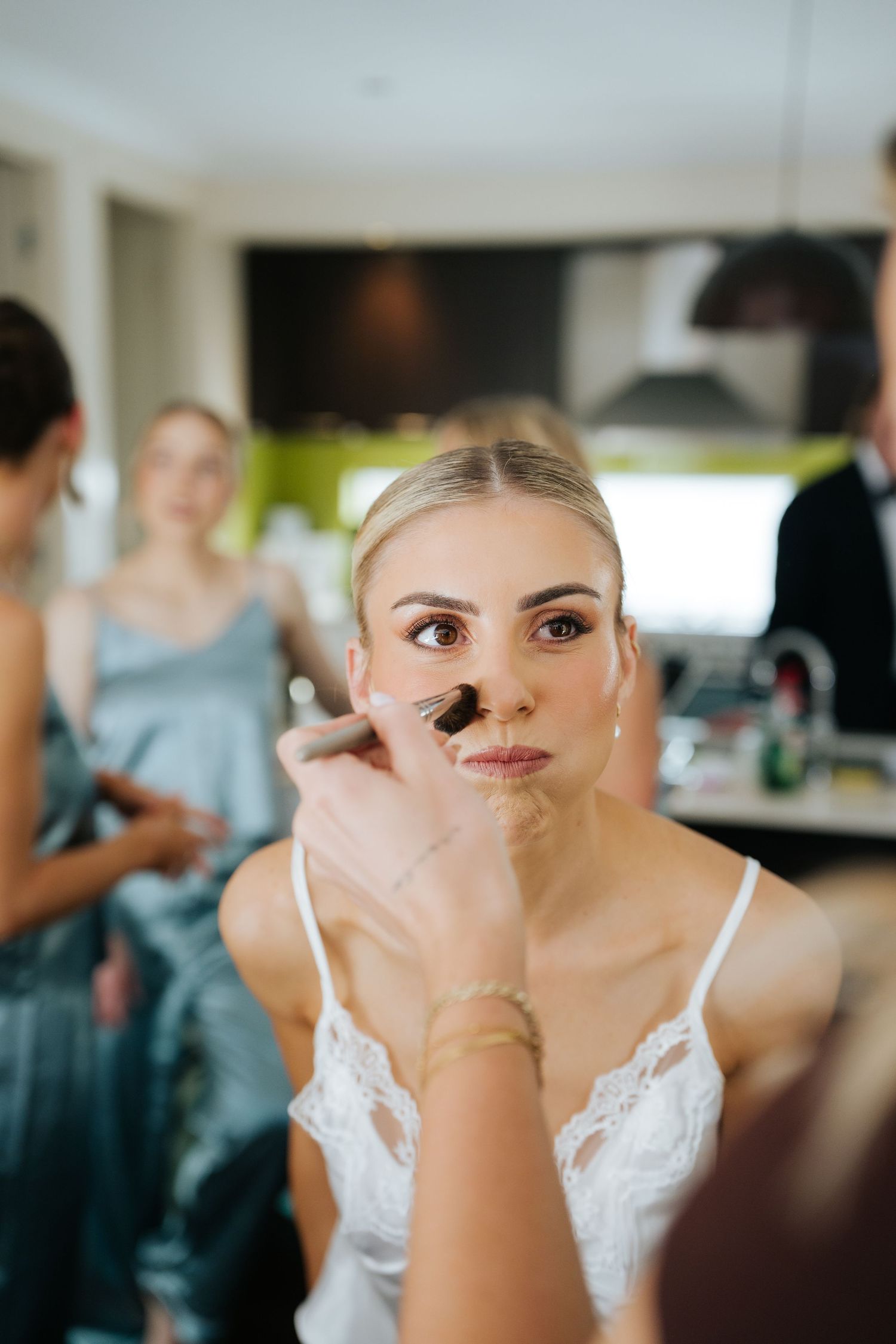 Professional makeup artist applying foundation to a blonde bride during wedding preparations indoors.