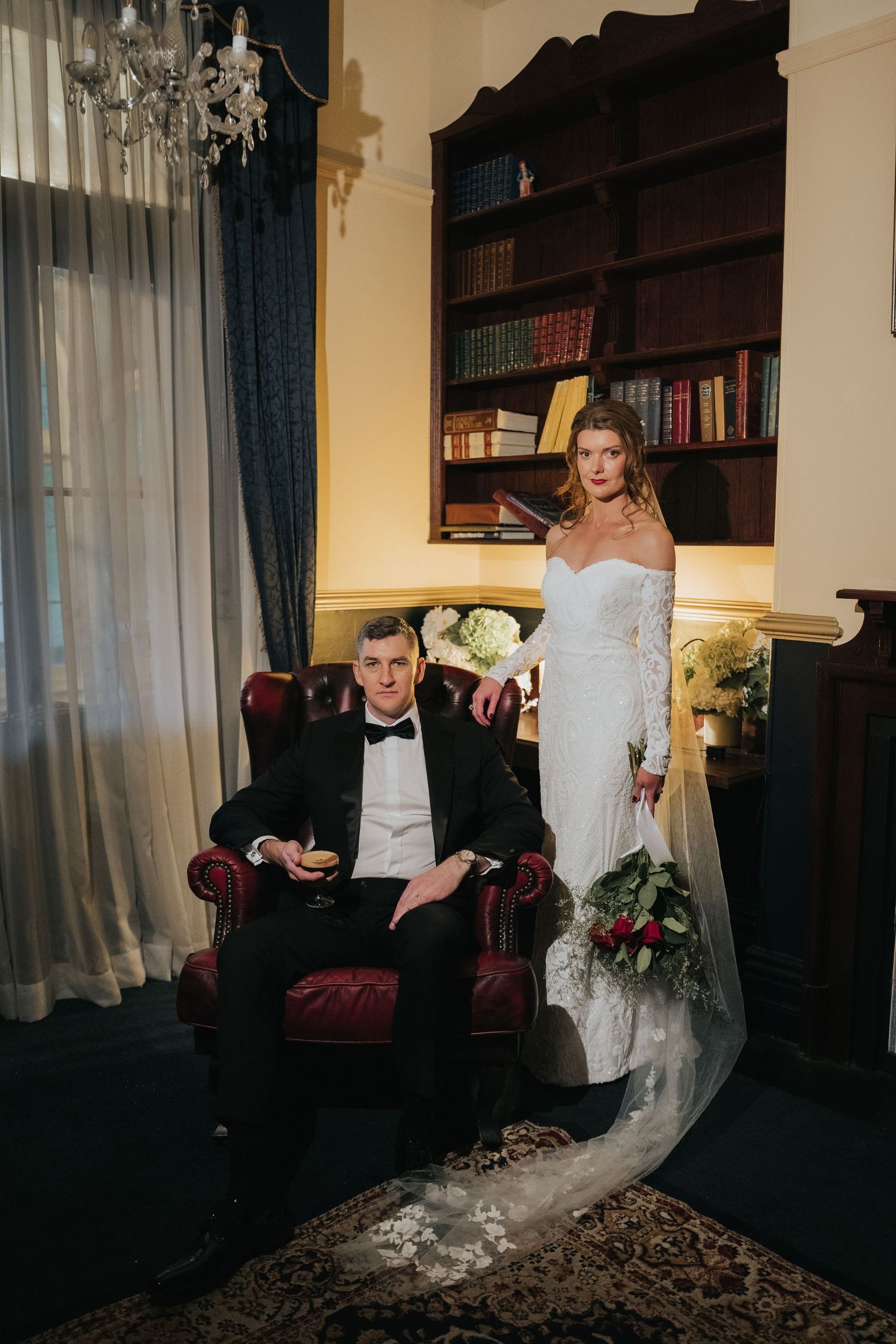 Elegant bride and groom posing in a classic library setting, bride in lace gown with veil, groom seated in red leather chair.