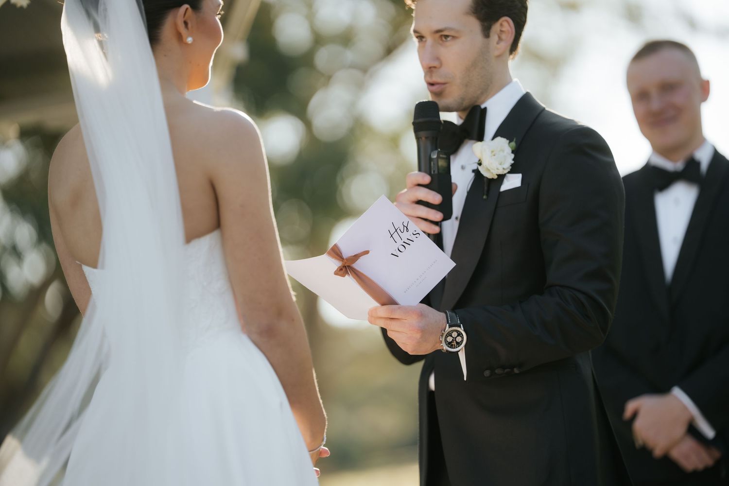 Groom in black tuxedo reading heartfelt vows from a card to his bride during outdoor wedding ceremony.