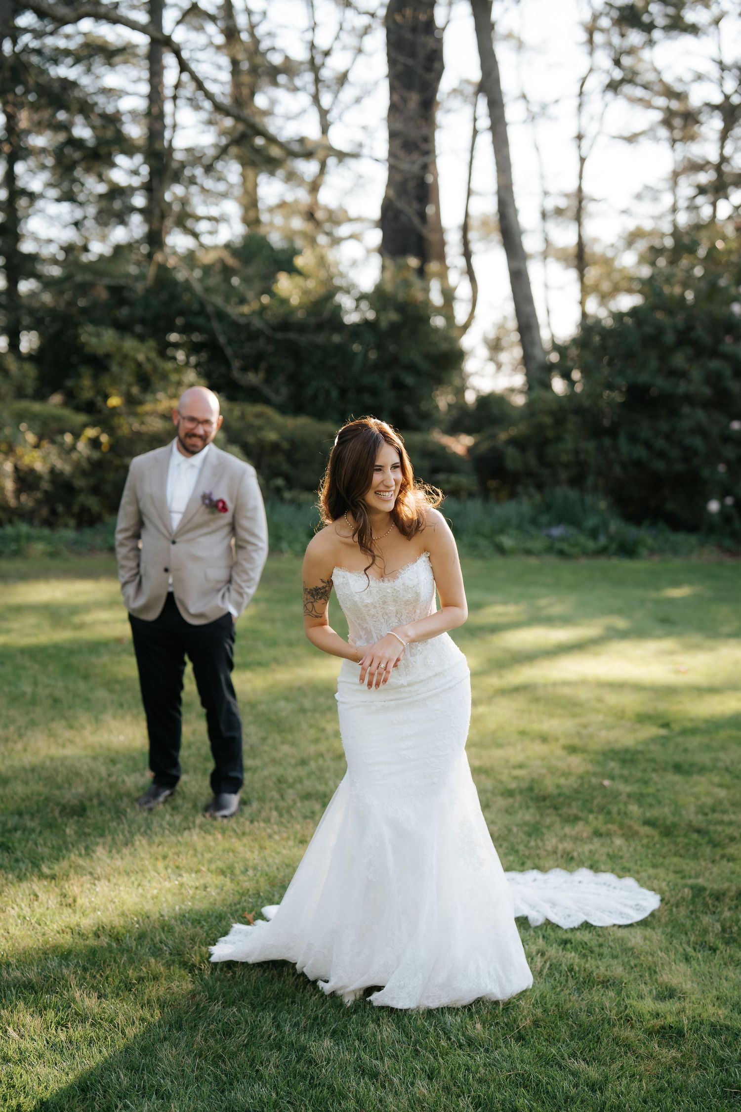 Bride in white mermaid gown smiles outdoors as groom in tan suit approaches on green lawn with trees behind.