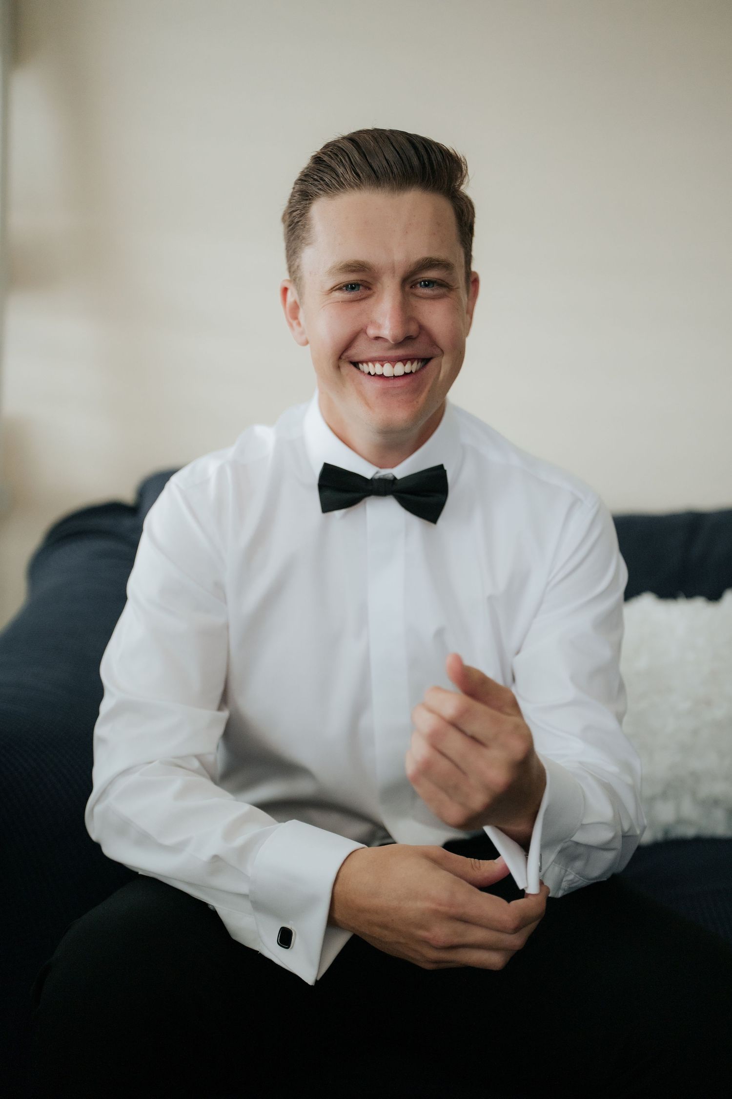 Smiling groom in white dress shirt and black bow tie adjusting cufflinks while seated on a dark sofa.