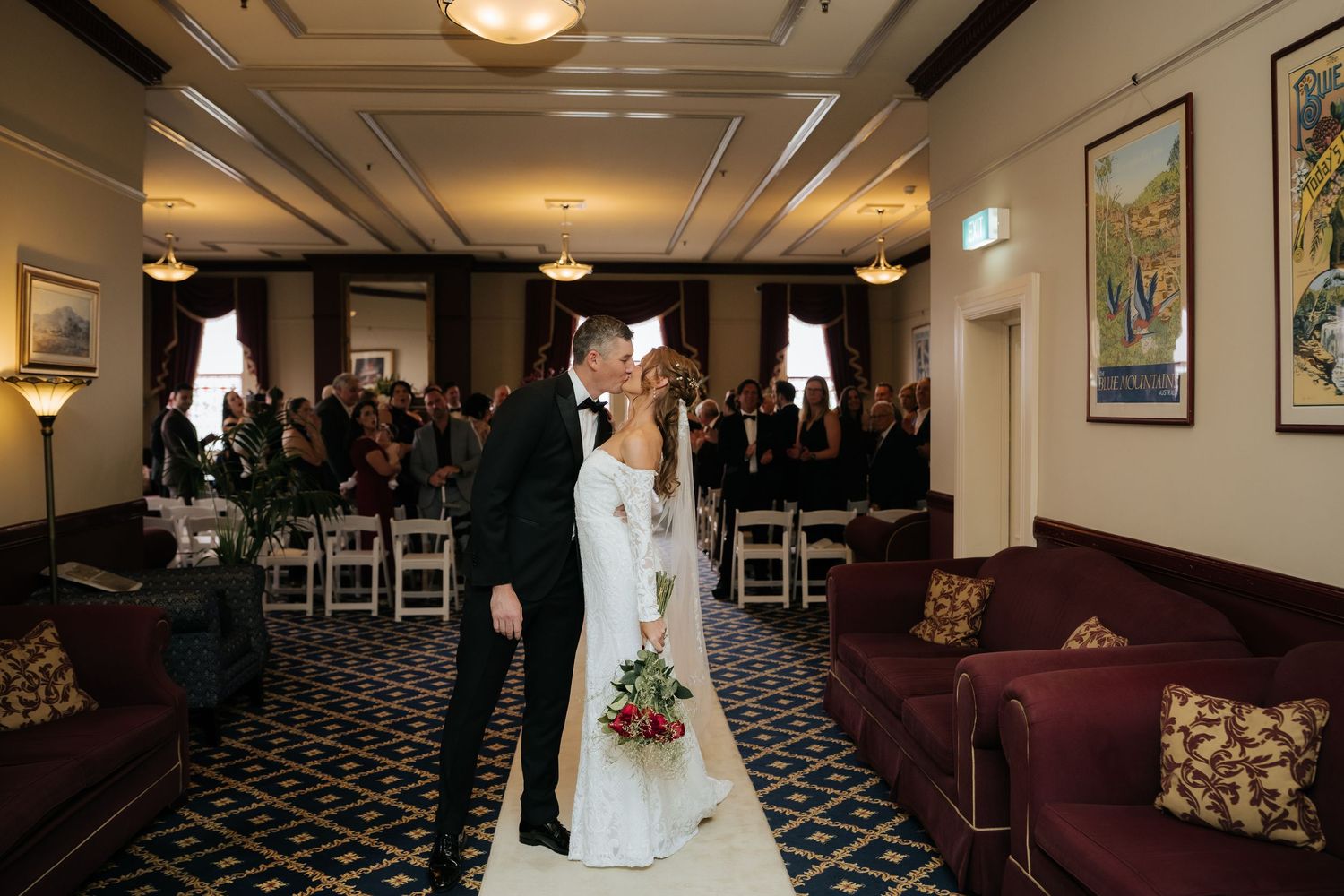Bride and groom share a kiss at indoor wedding ceremony in elegant venue with guests seated in background.