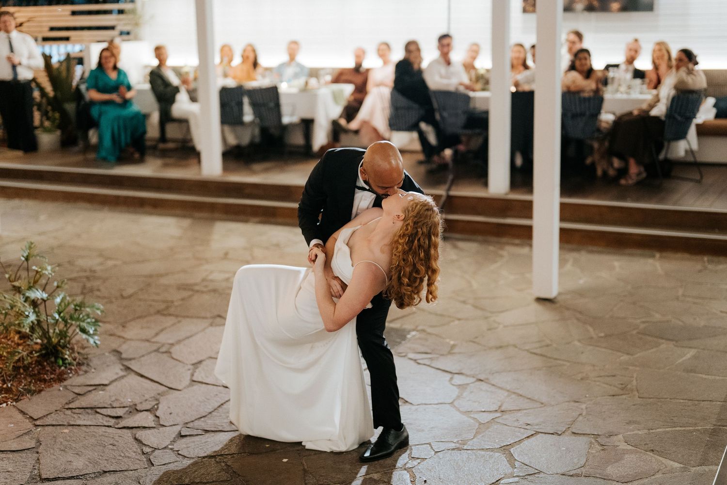 Groom dips bride during romantic first dance at elegant wedding reception with guests watching.