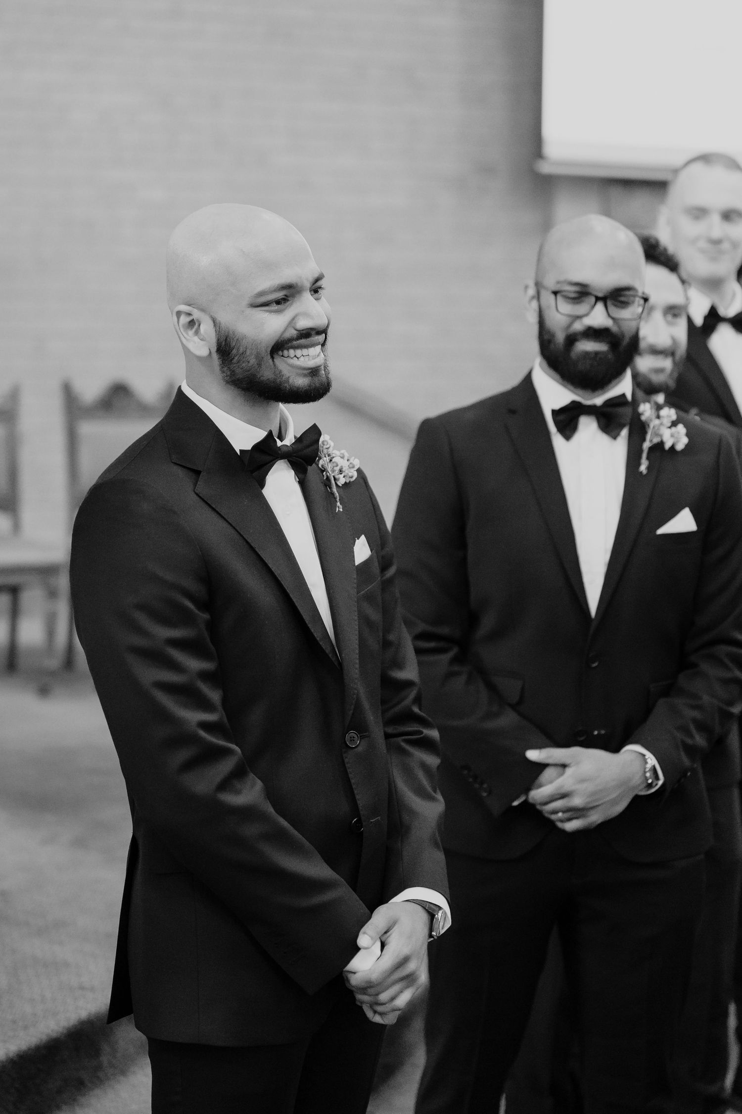 Black and white photo of two smiling grooms in tuxedos with boutonnieres at a wedding ceremony.