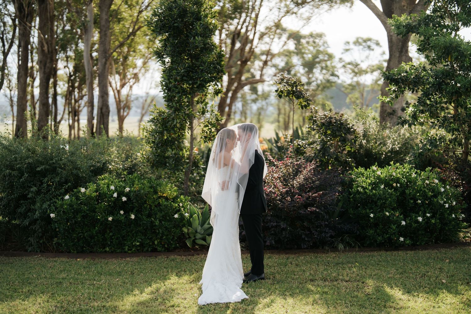 Bride and groom share a kiss under a flowing veil in a lush garden setting on their wedding day.