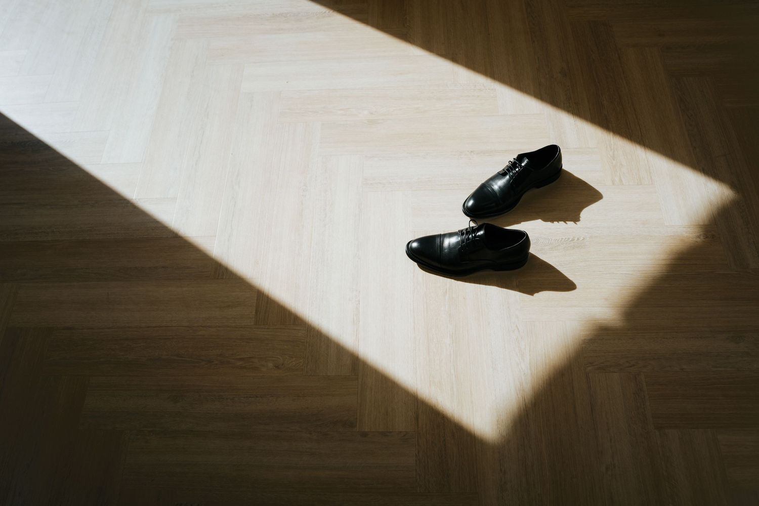 Black dress shoes resting on a herringbone wooden floor in a bright sunlit patch of light.