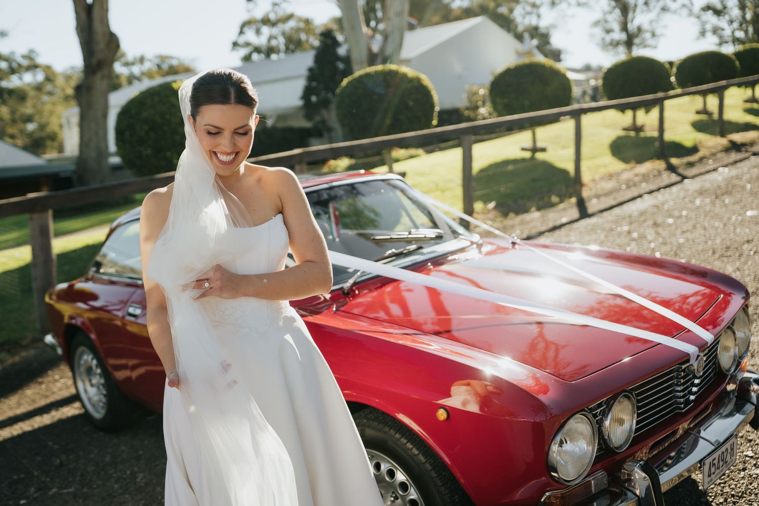 Laughing bride in white strapless gown and veil standing beside a classic red vintage car on wedding day.