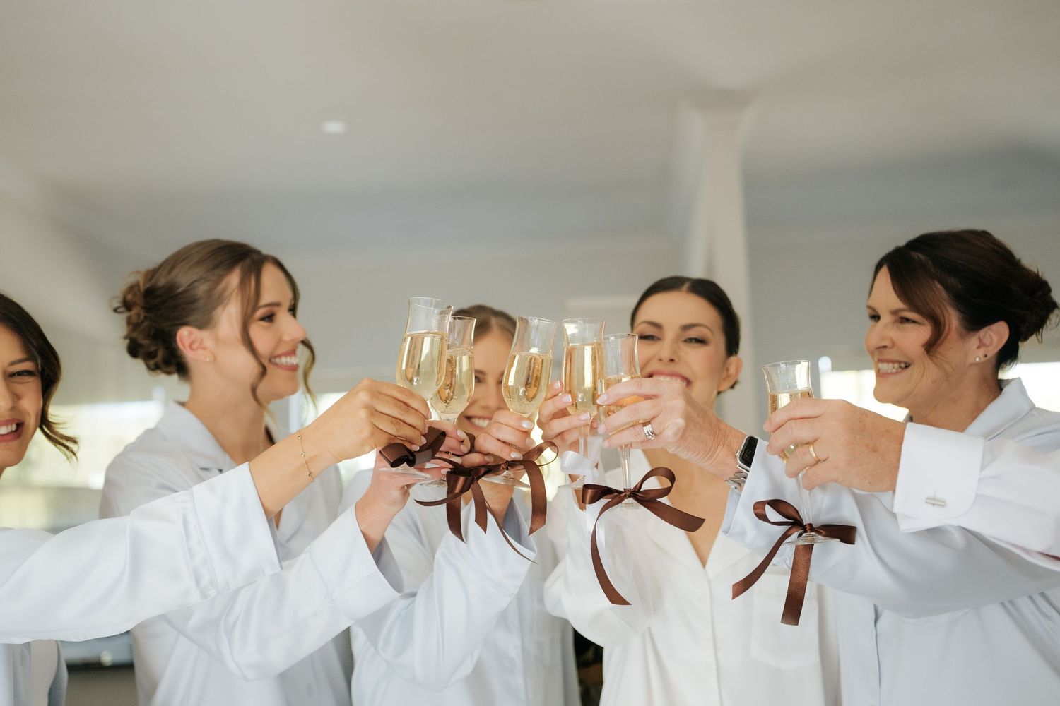 Bridesmaids in white robes with brown bows toasting with champagne glasses during bridal party preparations.