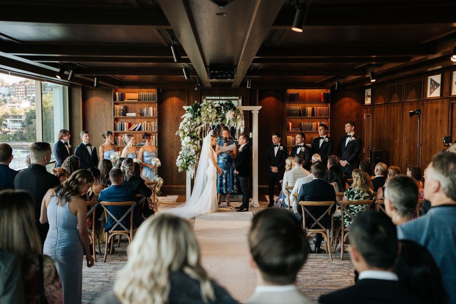 Elegant indoor wedding ceremony in wood-paneled venue with floral arch and seated guests.