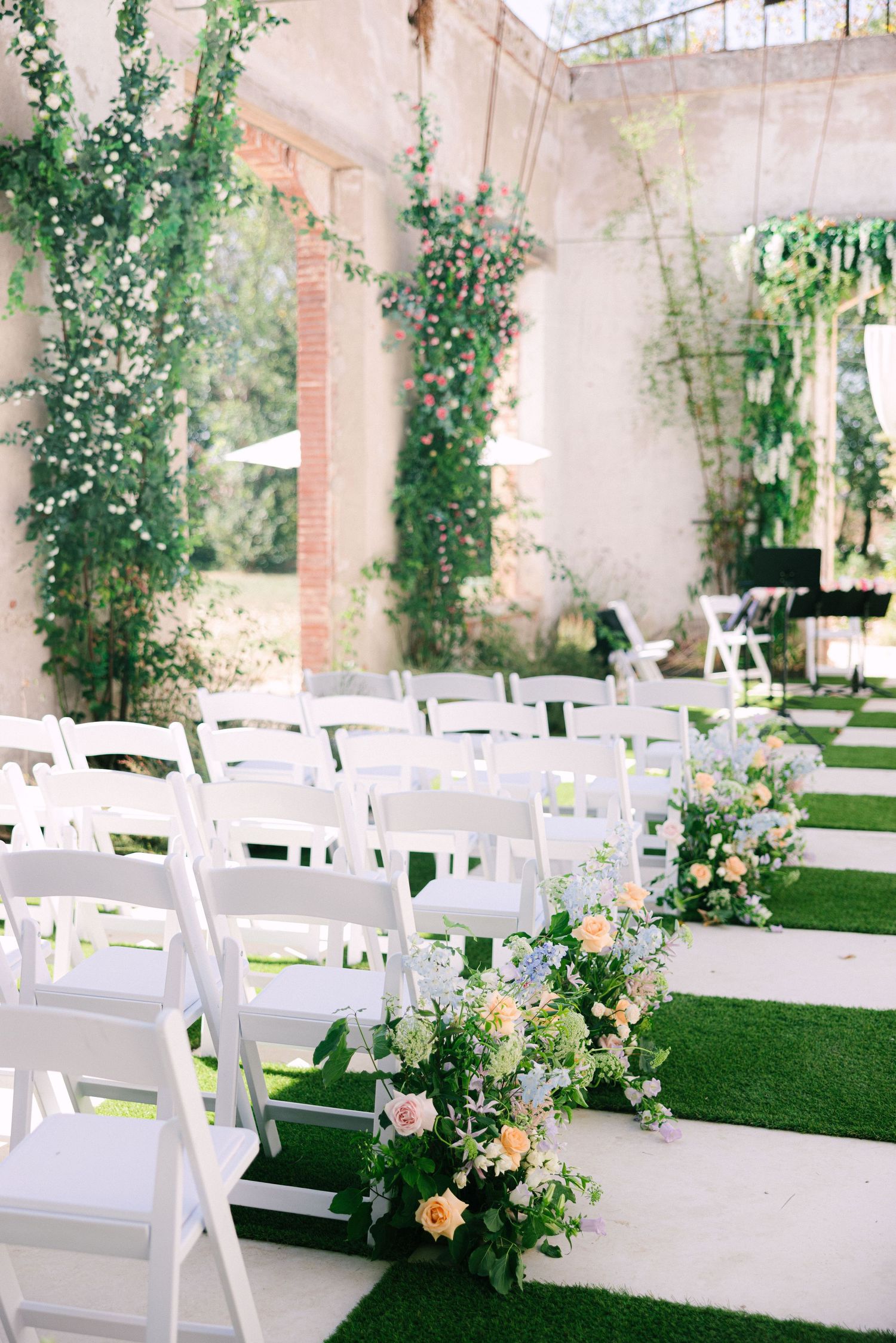 Elegant outdoor wedding ceremony setup with white folding chairs and romantic floral arrangements along a covered walkway.