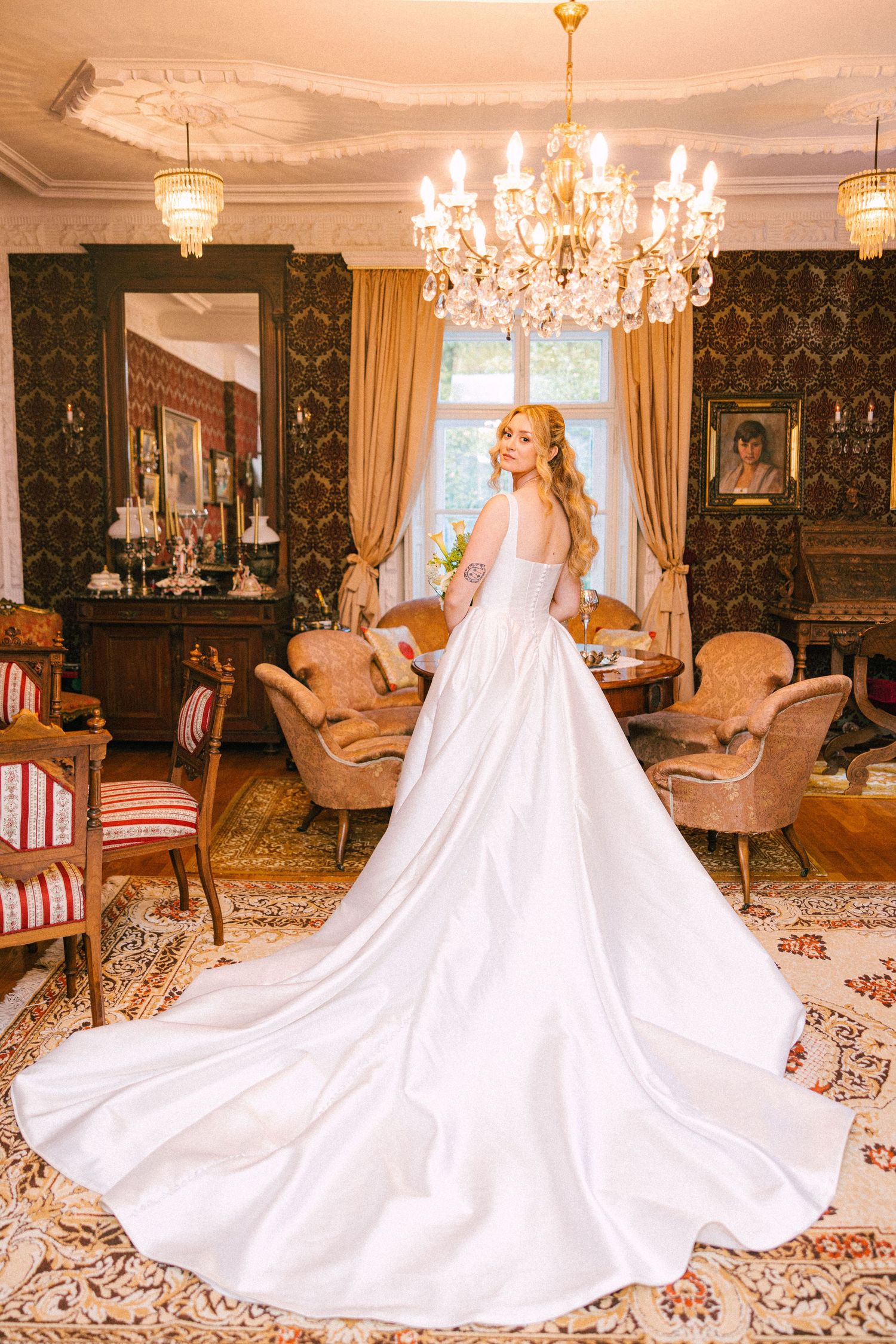 A bride in a flowing white wedding gown with a dramatic cathedral train poses in an elegant room with chandeliers.
