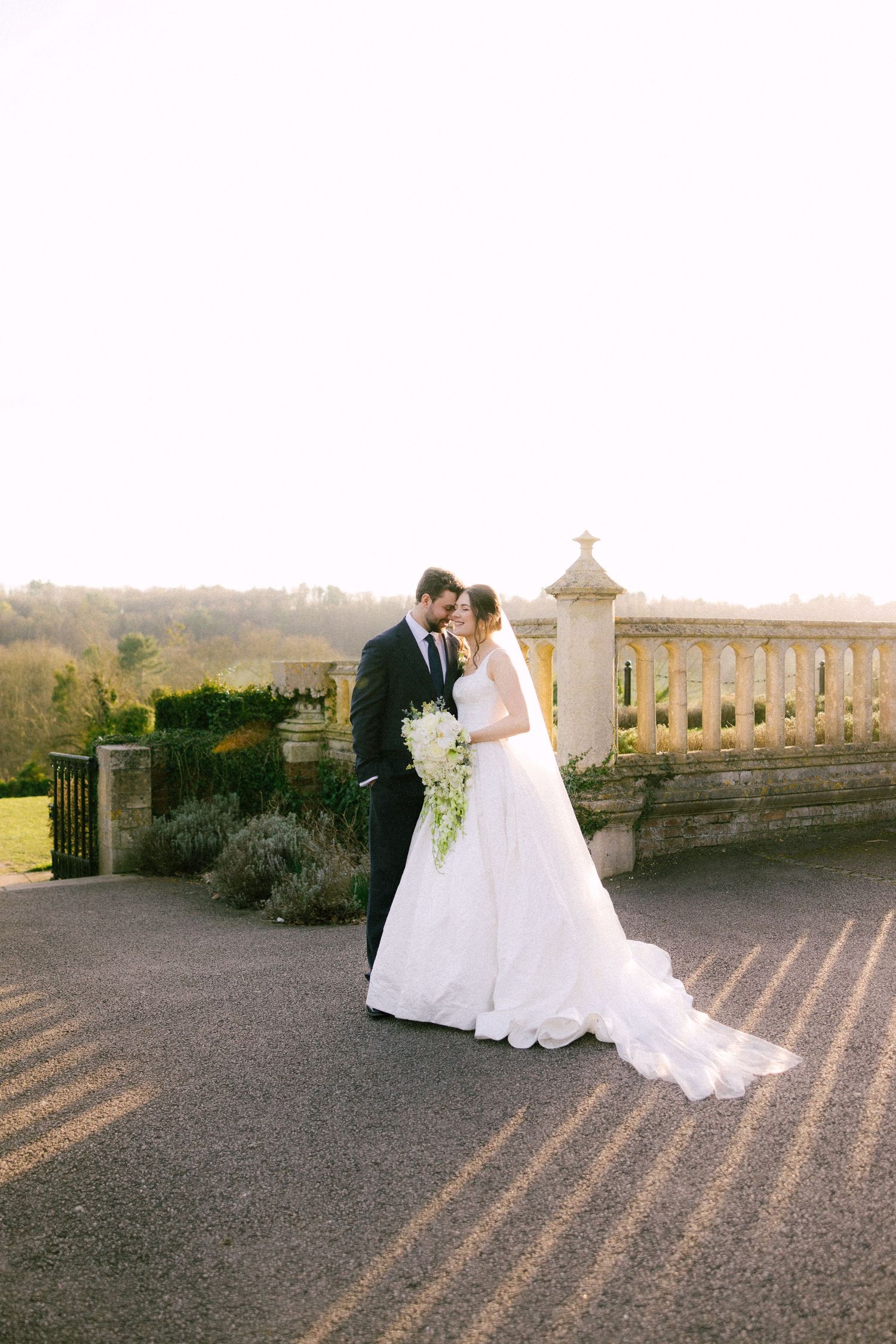 A couple in wedding attire embraces on a stone terrace during sunset, creating long dramatic shadows.