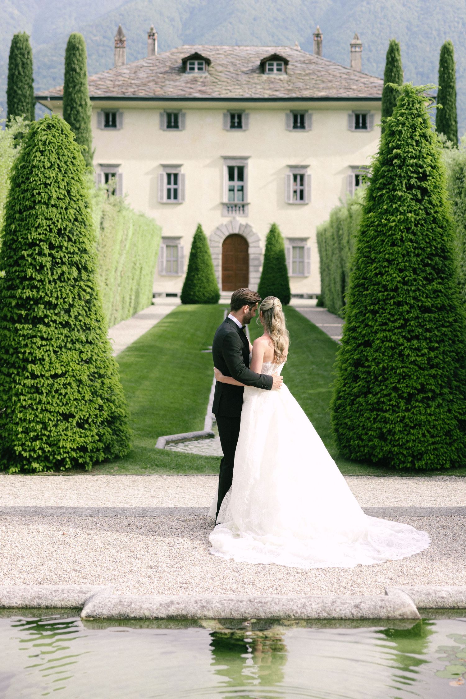 A couple in wedding attire stands on a gravel path lined with manicured cypress trees at an elegant Italian villa.