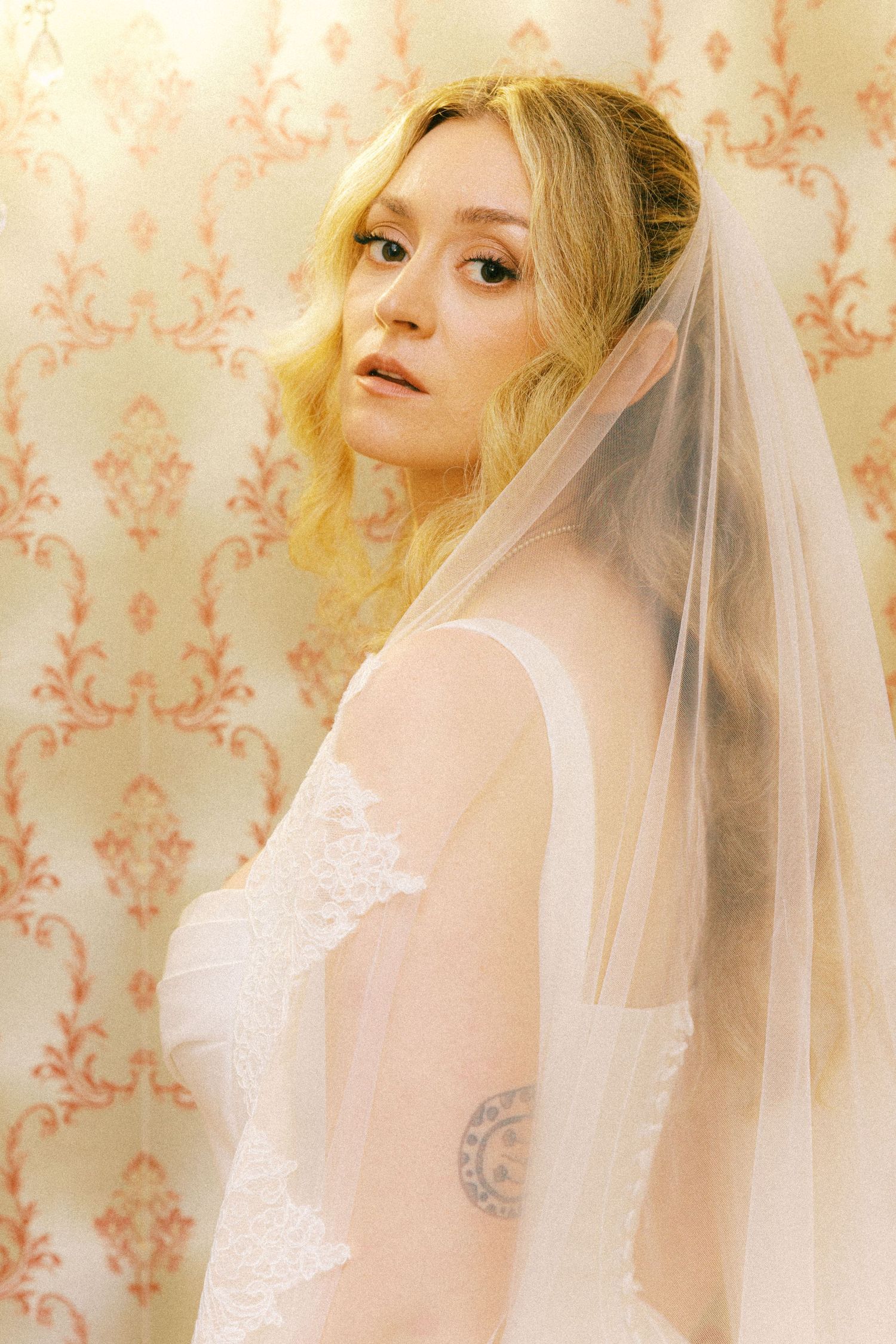 Ethereal bridal portrait with soft veil against peach damask wallpaper in dreamy vintage style lighting.