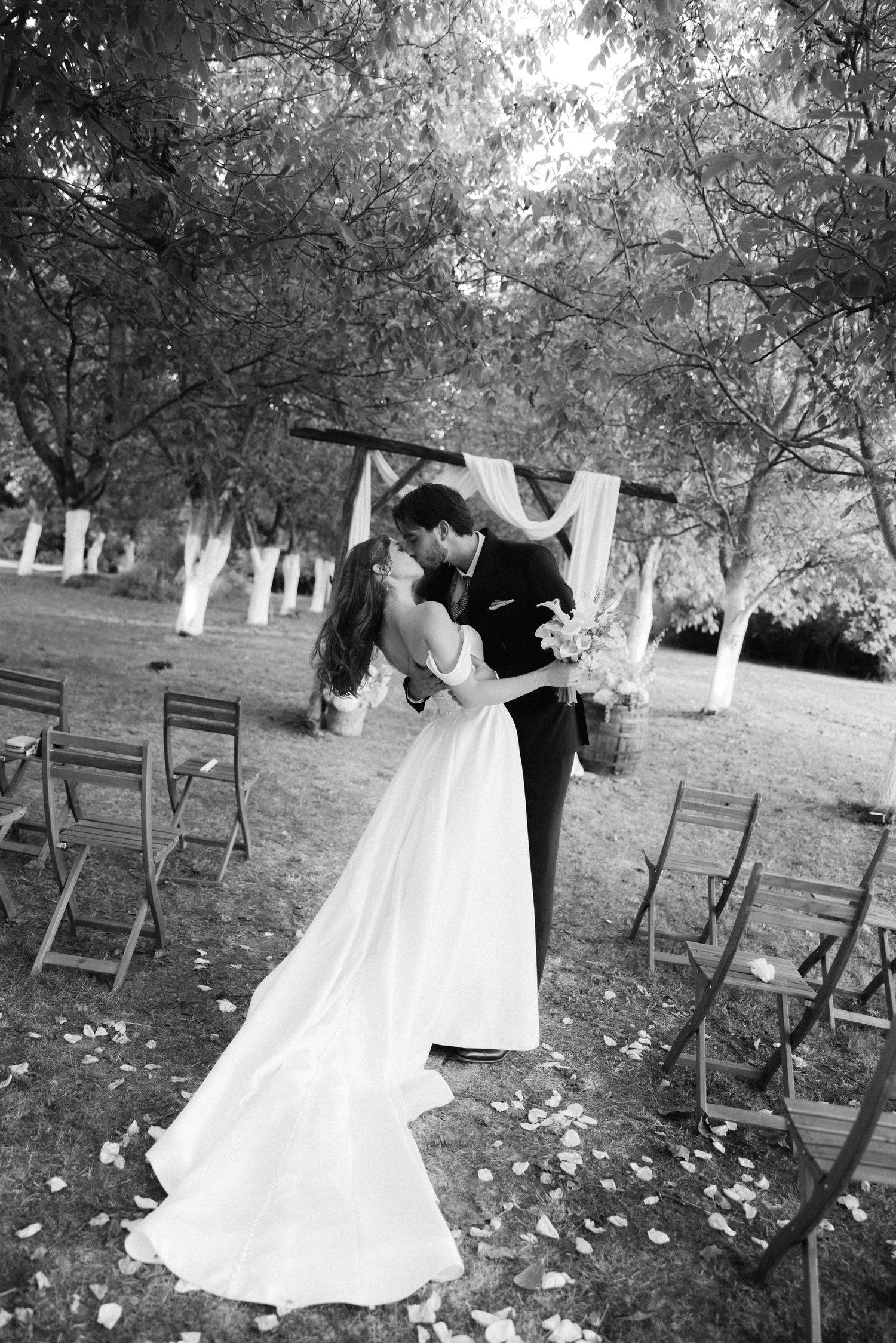 Black and white wedding photo series showing a couple dipping and dancing outdoors among wooden chairs and fallen leaves.