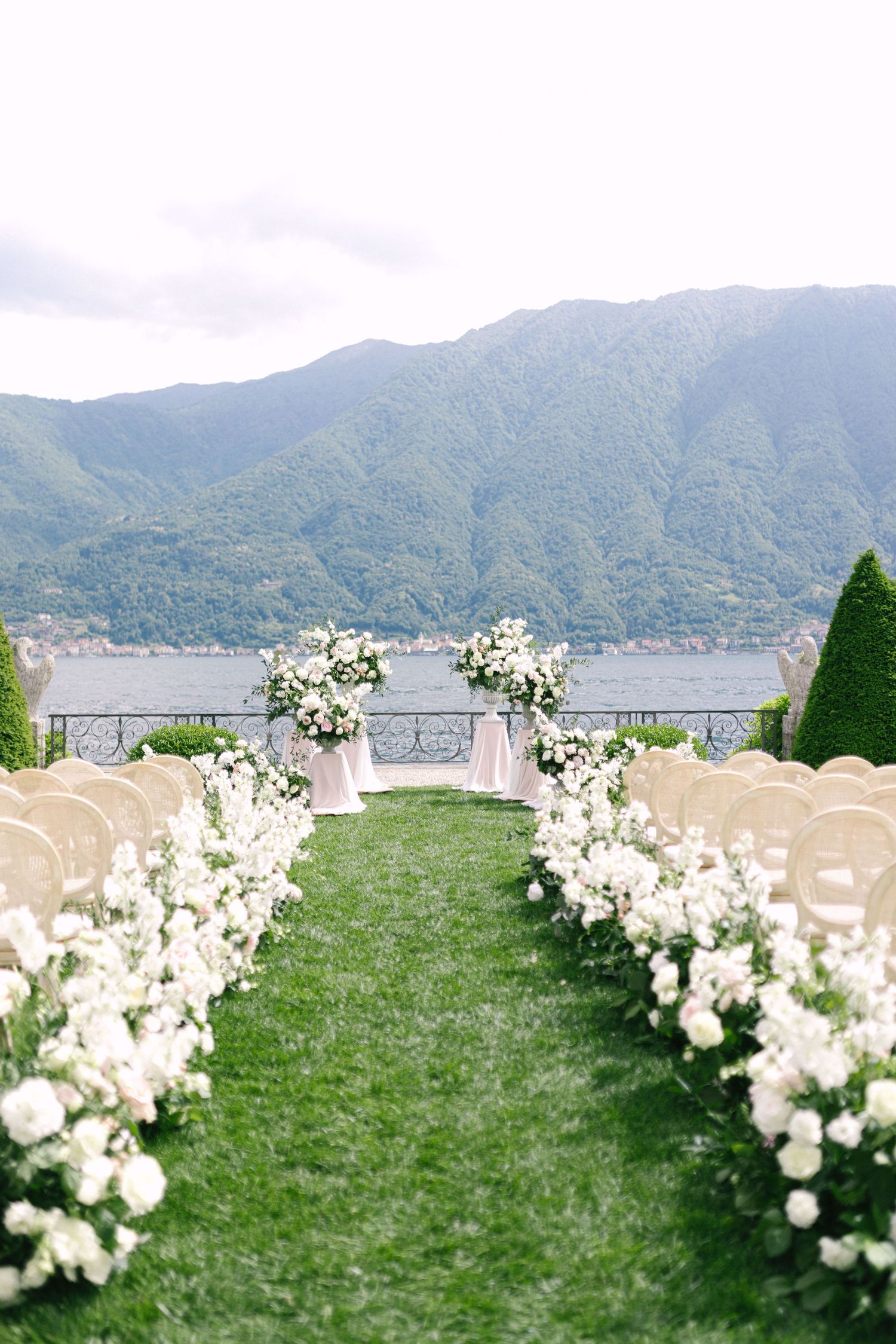 White floral arrangements line a grassy outdoor ceremony space overlooking a mountain lake landscape.