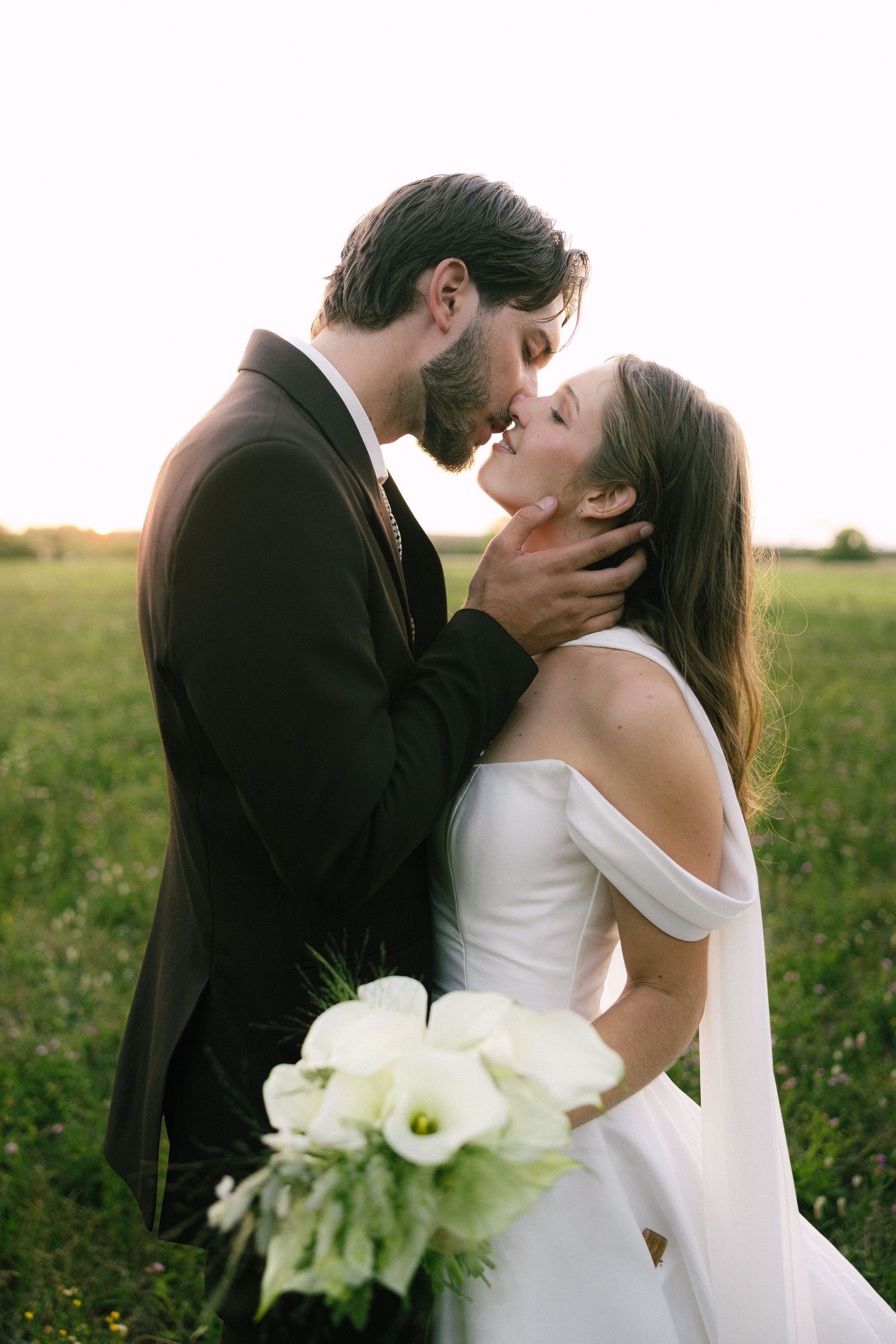 Romantic wedding couple embraces in golden field at sunset with white calla lily bouquet.